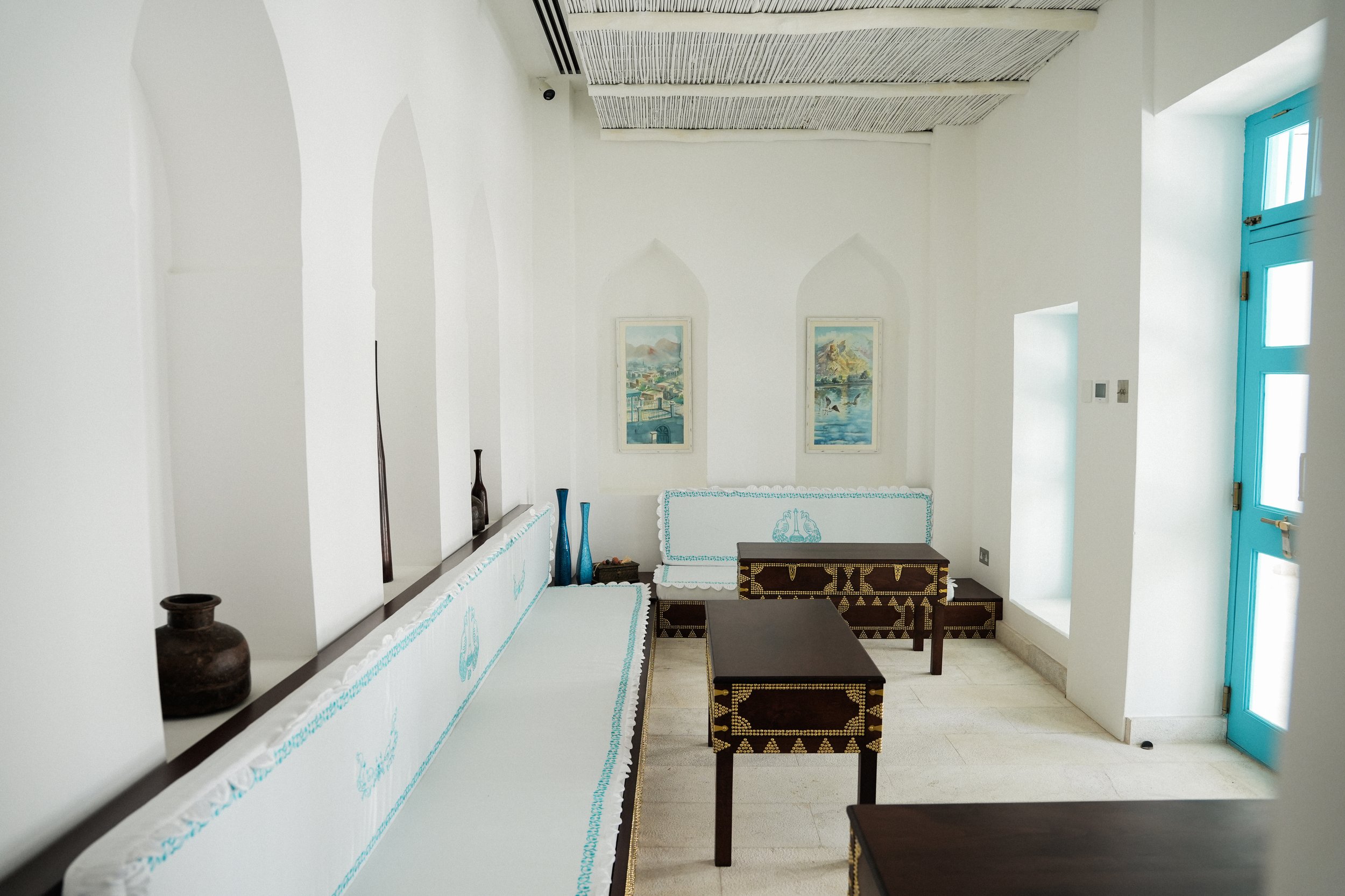 Interior of the sebla with white walls, decorated with framed landscape paintings, blue accents on a door and furniture, and various decorative vases and boxes.