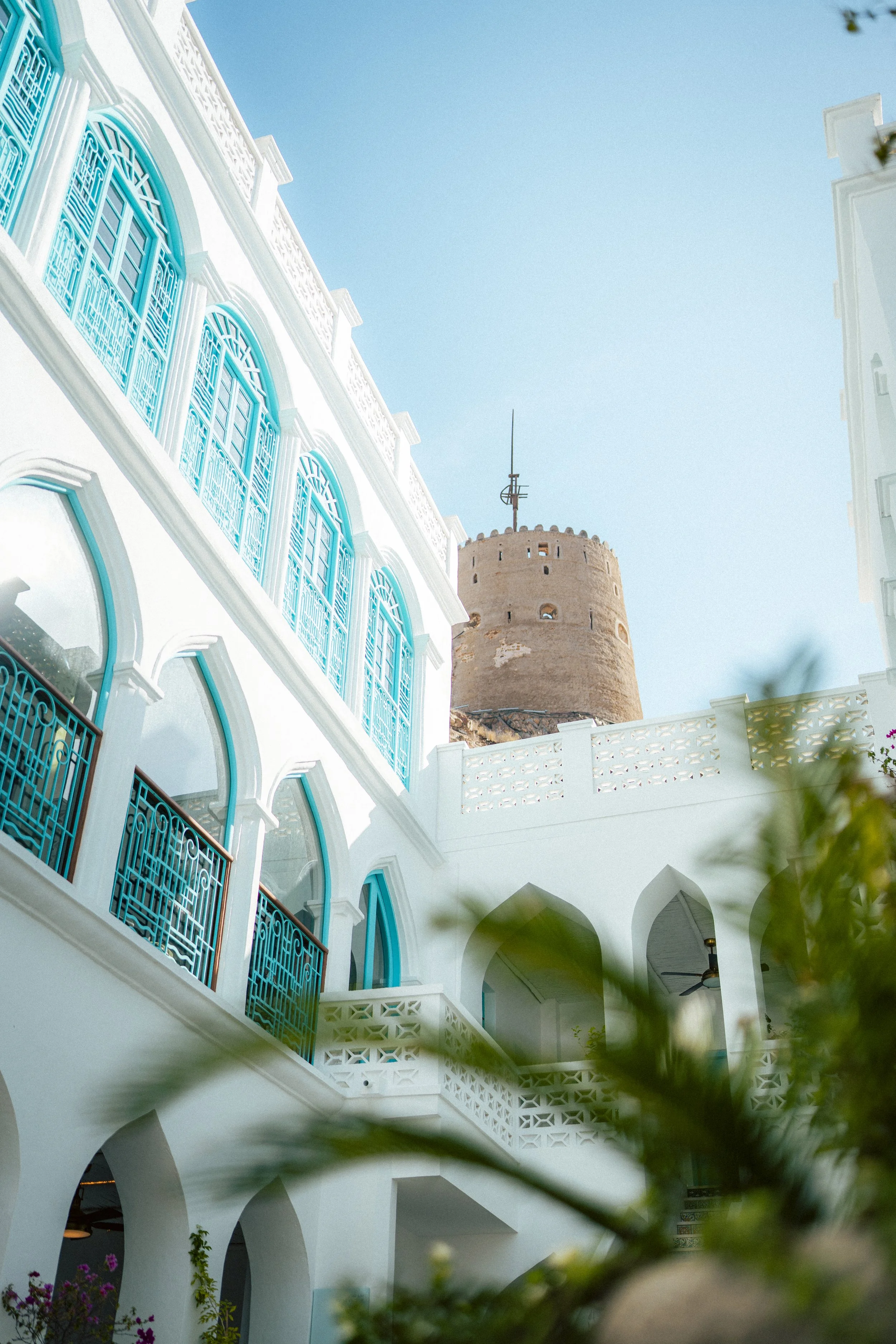 A white building with blue window frames and decorative ironwork,Muttrah fort in the background, and clear blue sky.