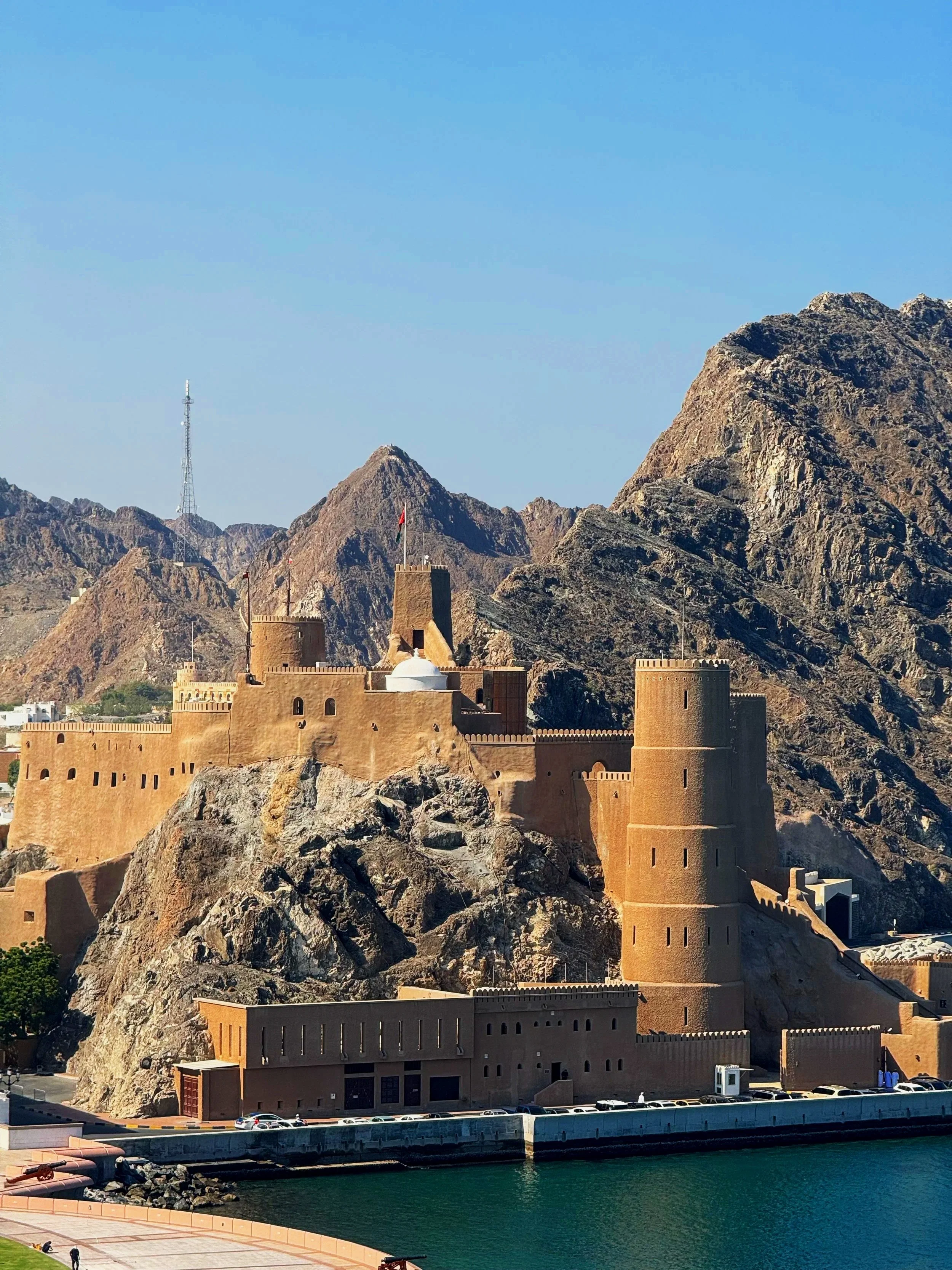 A historic desert fortress with tall watchtowers and thick walls, situated on rocky terrain near water, with mountains in the background and a clear blue sky overhead.
