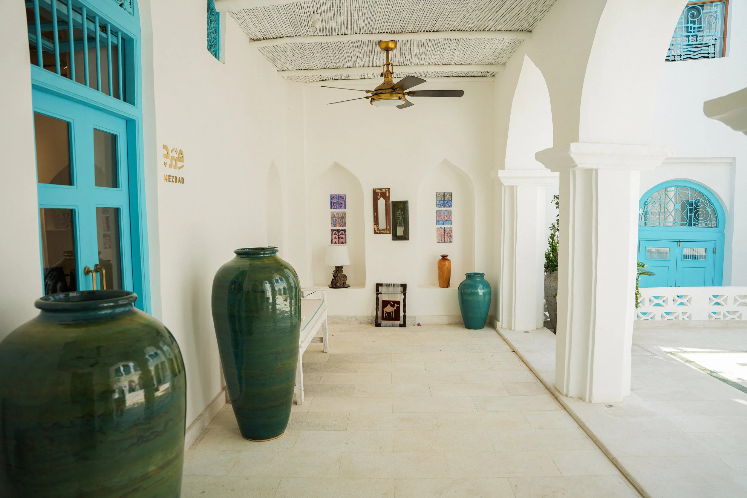 A white-walled interior with blue doors and window frames, decorated with various colorful framed artwork, large green vases, a small table, and a ceiling fan, with architectural arches and a view outside.