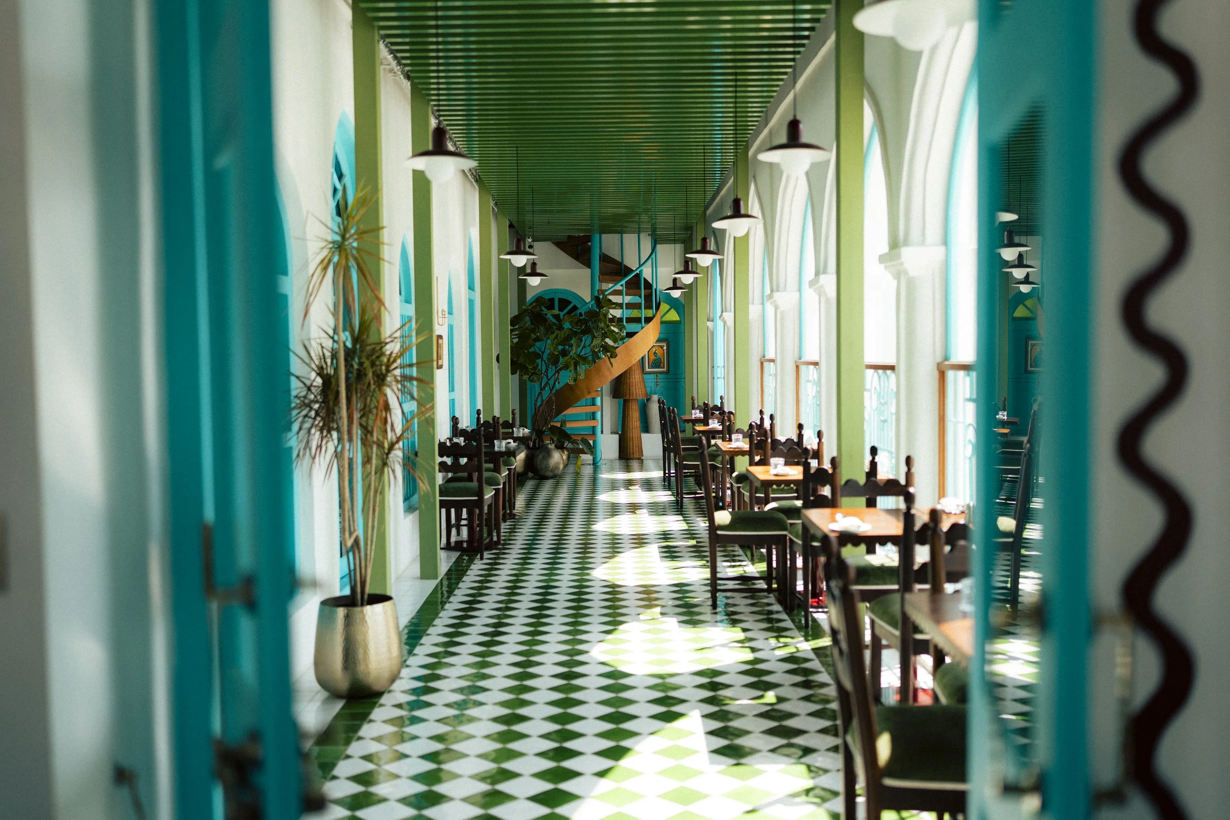 Interior of a restaurant with green and white checkered floor, wooden tables with chairs, large windows with arched frames, hanging pendant lights, and potted plants, viewed through a teal-colored doorway.