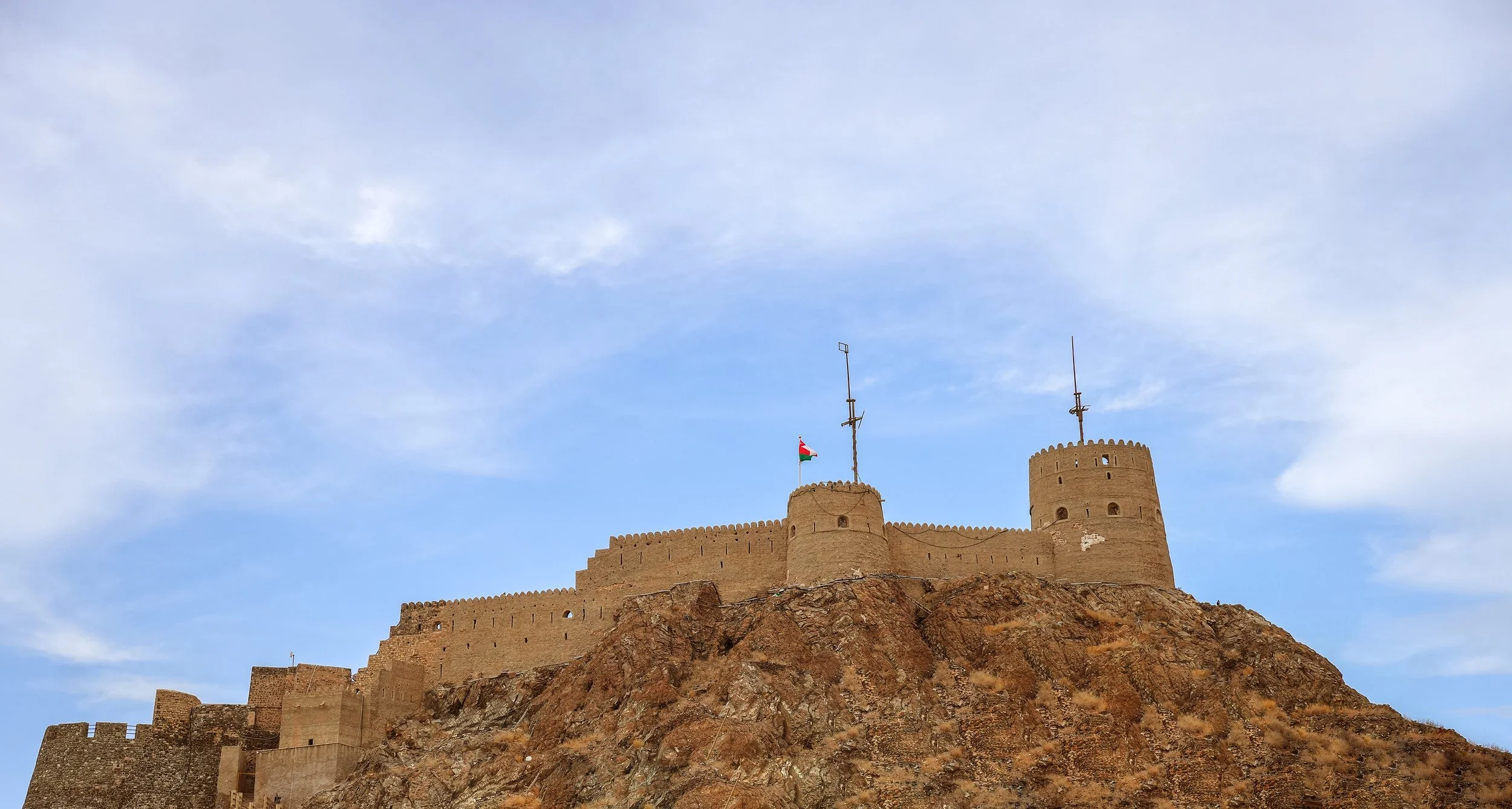 Ancient desert fortress with stone walls and towers, flying a flag, under a partly cloudy sky.