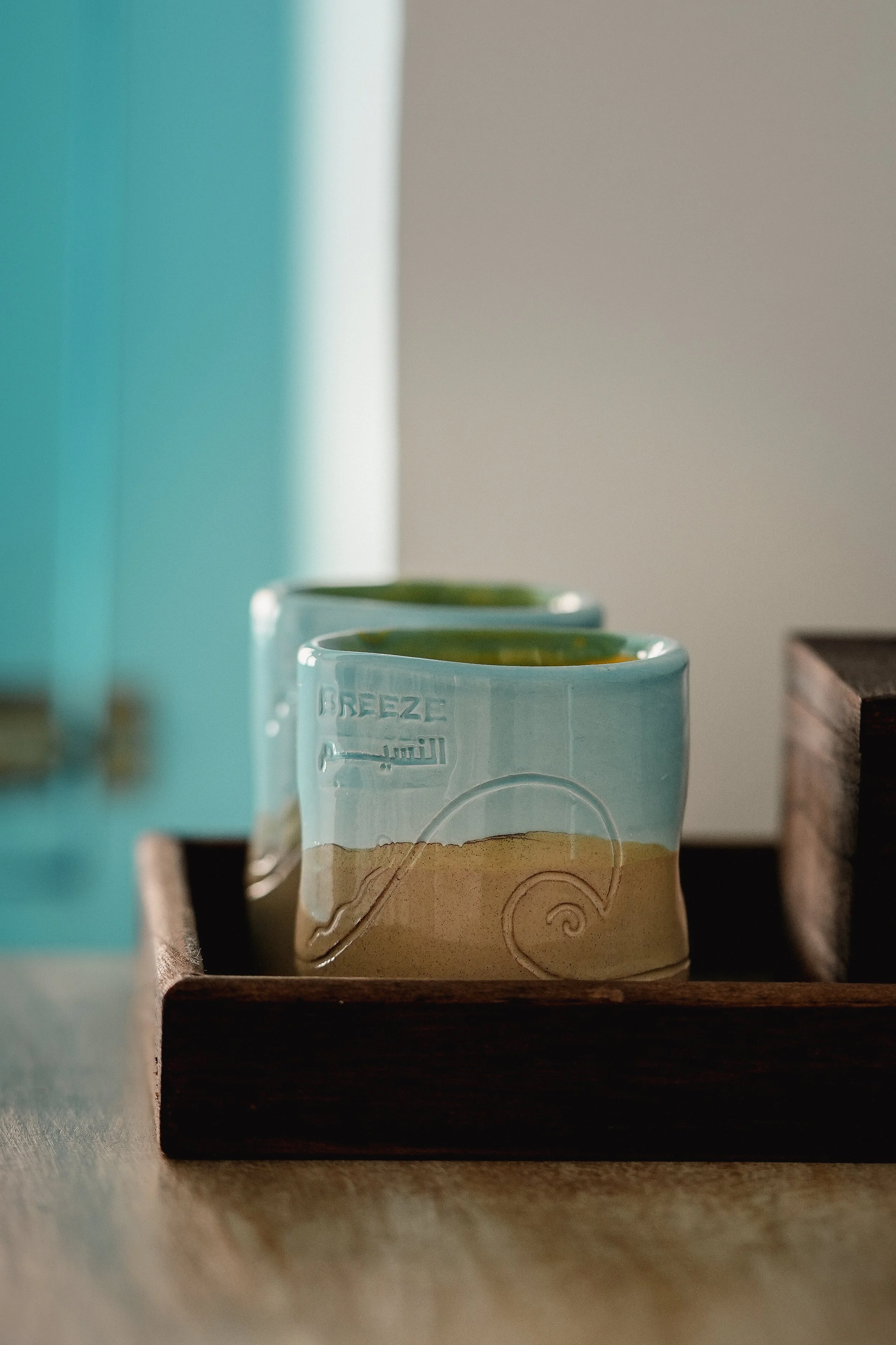Close-up of a beige and green tea cup with the word 'Breeze' embossed on its side, placed on a dark wooden tray on a light-colored surface from the rooms name.