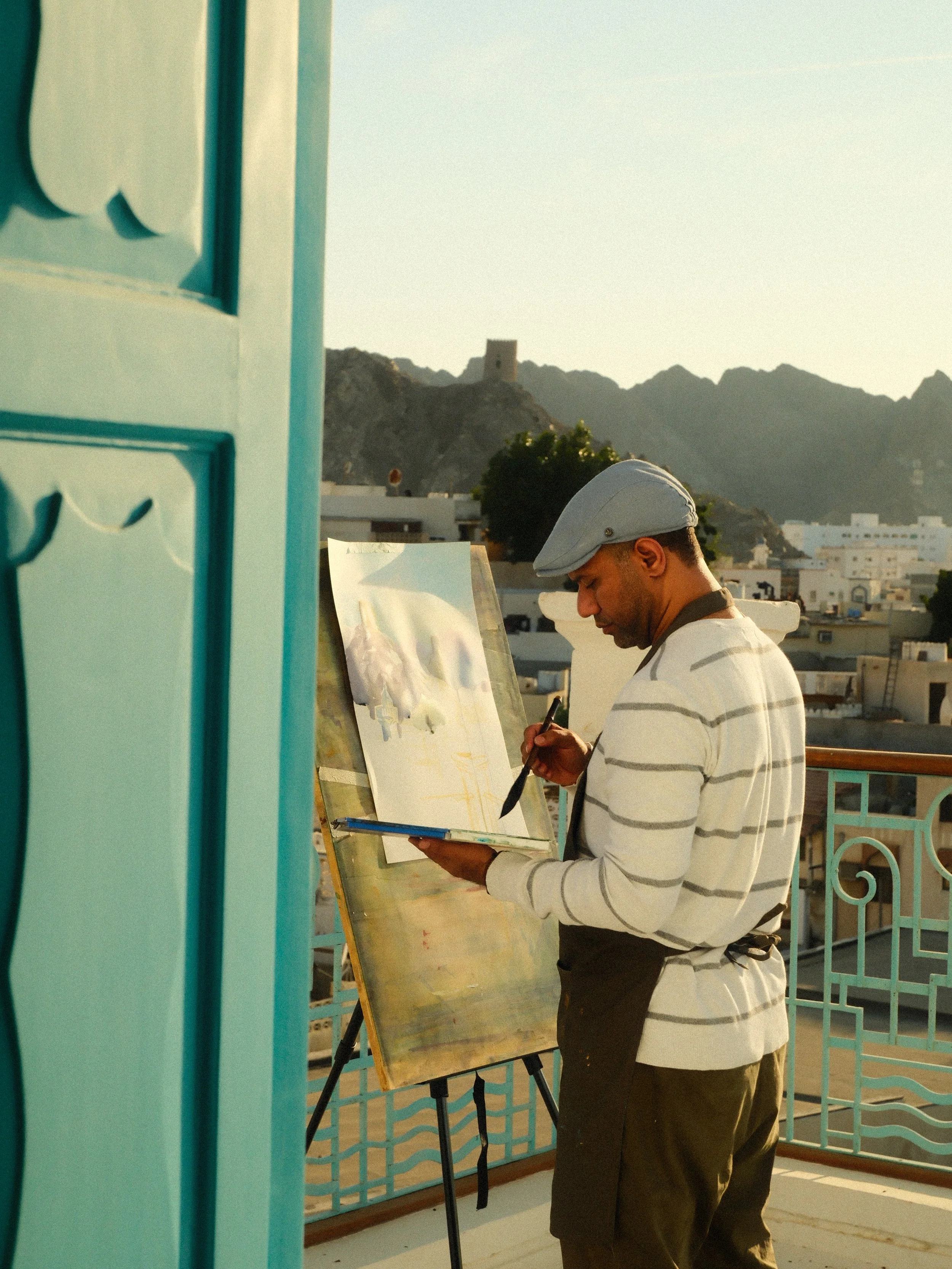 A man wearing a gray flat cap and a striped sweater paints on a canvas set up on an easel outdoors during sunset, with a mountain landscape and white buildings in the background.