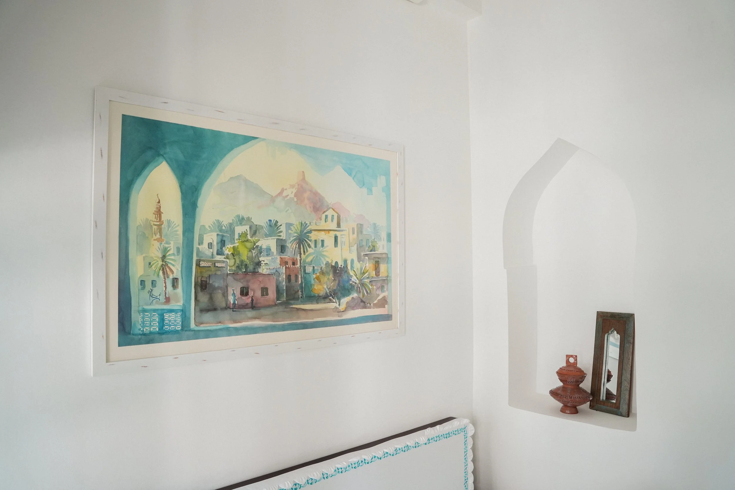 Interior wall with a colorful watercolor painting of a town scene, a decorative jar, and a mirror on a shelf.