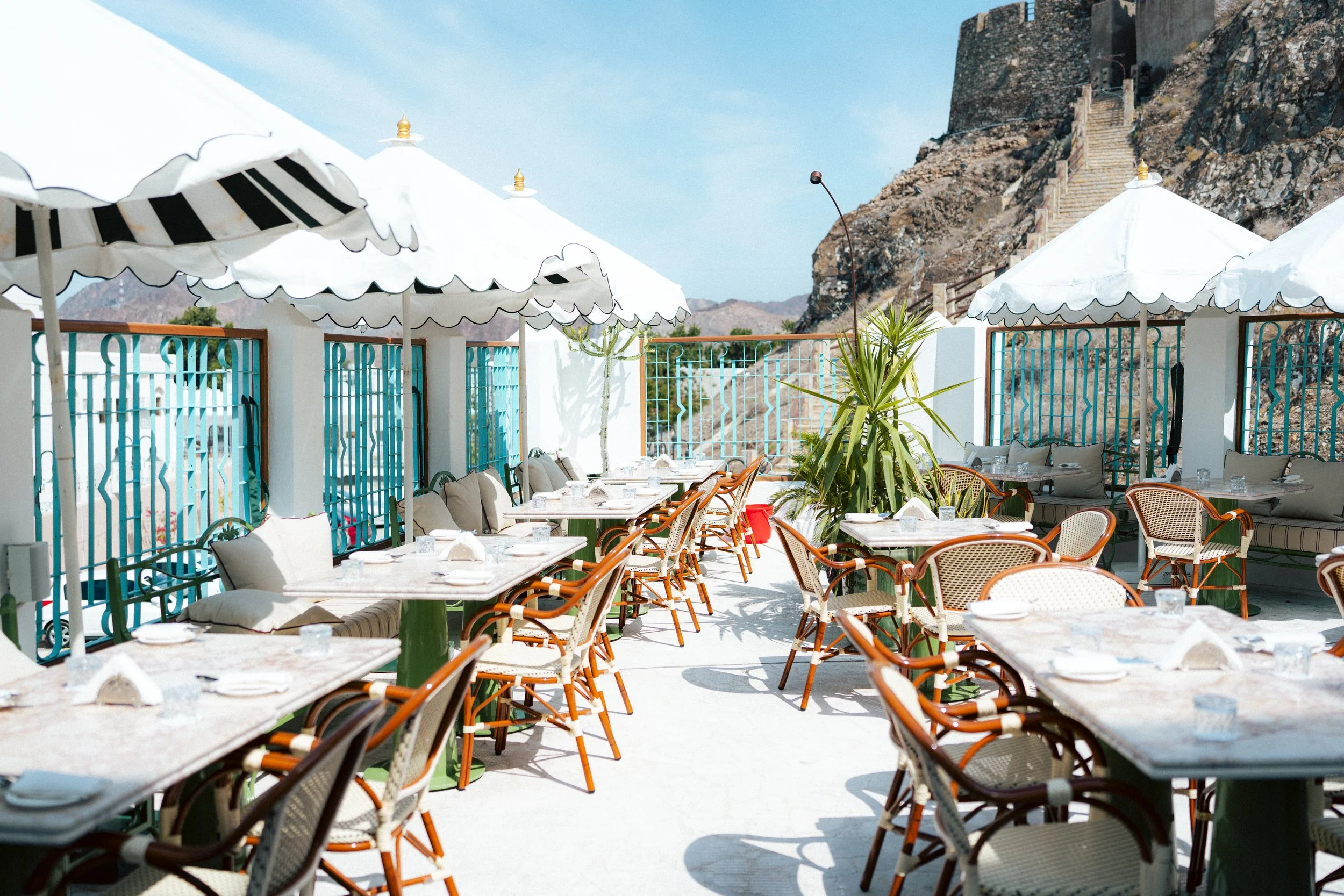Outdoor restaurant seating with tables, chairs, umbrellas, and a mountain staircase in the background.
