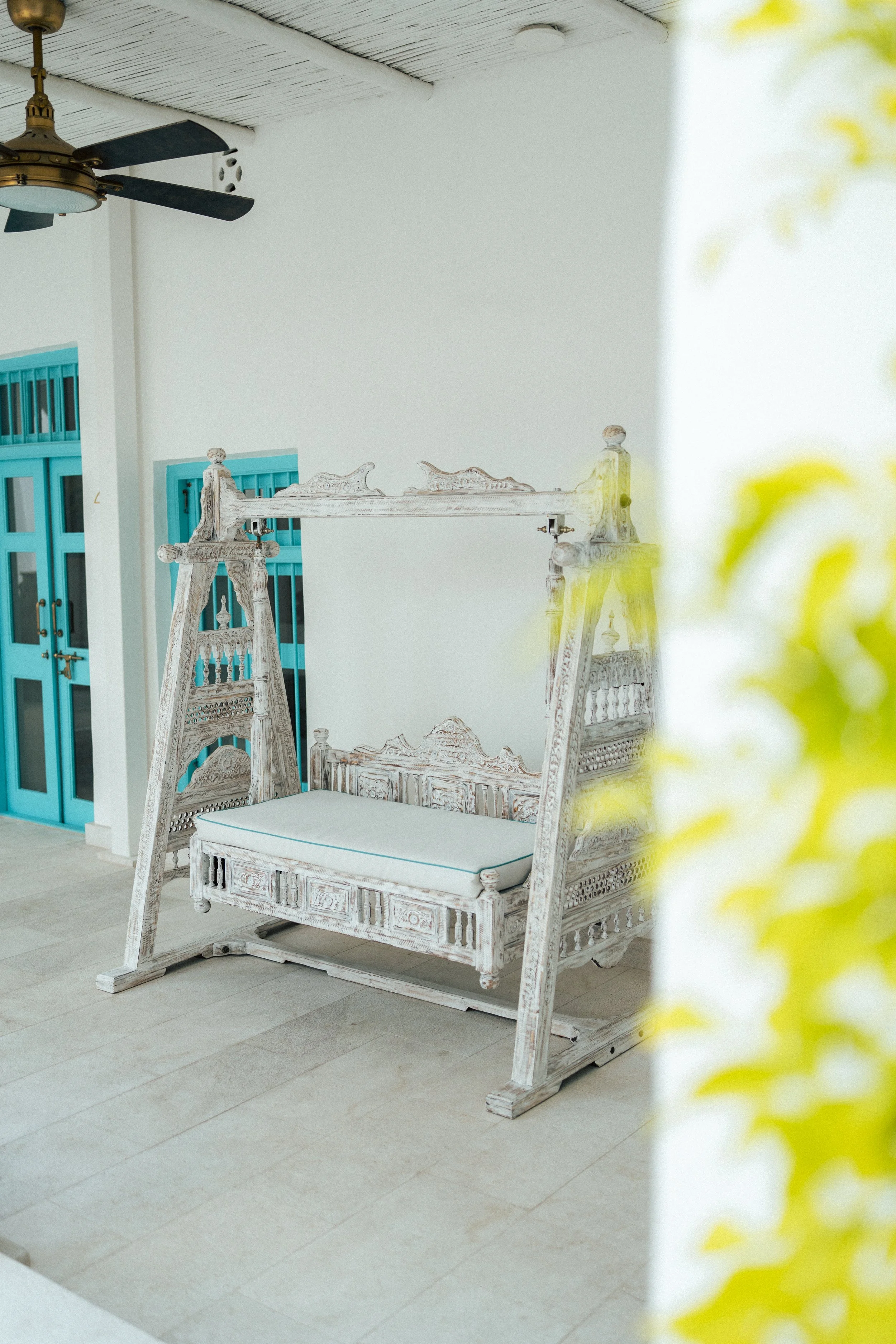A white wooden outdoor sofa with intricate carvings, positioned on beige tiles, with turquoise doors and a ceiling fan with dark blades overhead on a white ceiling, and some out-of-focus yellow flowers on the right side.