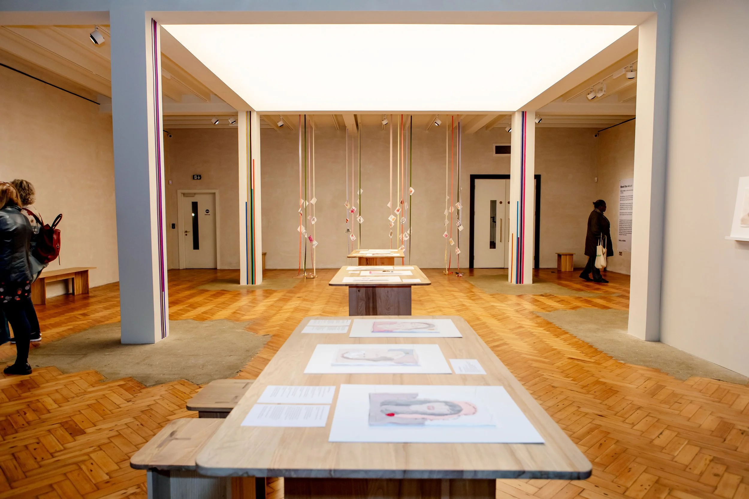 An art gallery with wooden floors and beige walls, displaying colorful hanging sculptures and artwork on tables.