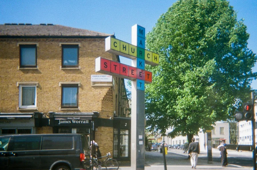 Street sign spelling 'Market', 'Church', and 'Street' in colorful blocks with a brick building, trees, and pedestrians in the background.