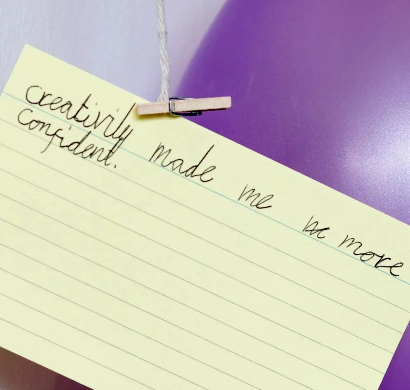 A yellow lined note card clipped to a purple surface with a small clothespin, containing handwritten text that reads 'creativity made me confident.'