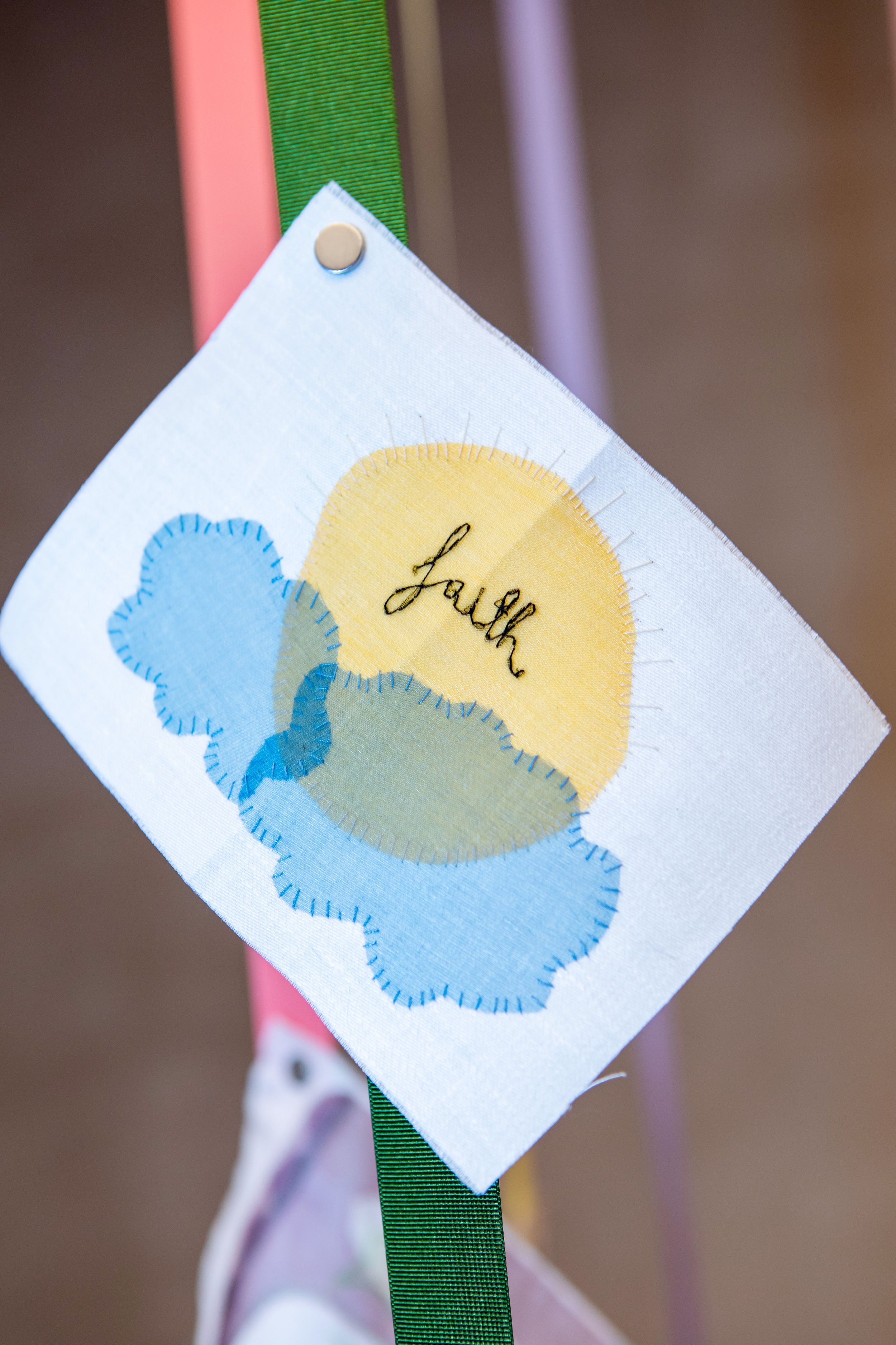 A stitched cloth decoration with a yellow sun, blue and green clouds, and the embroidered word 'Laugh' in black, attached to a green ribbon.