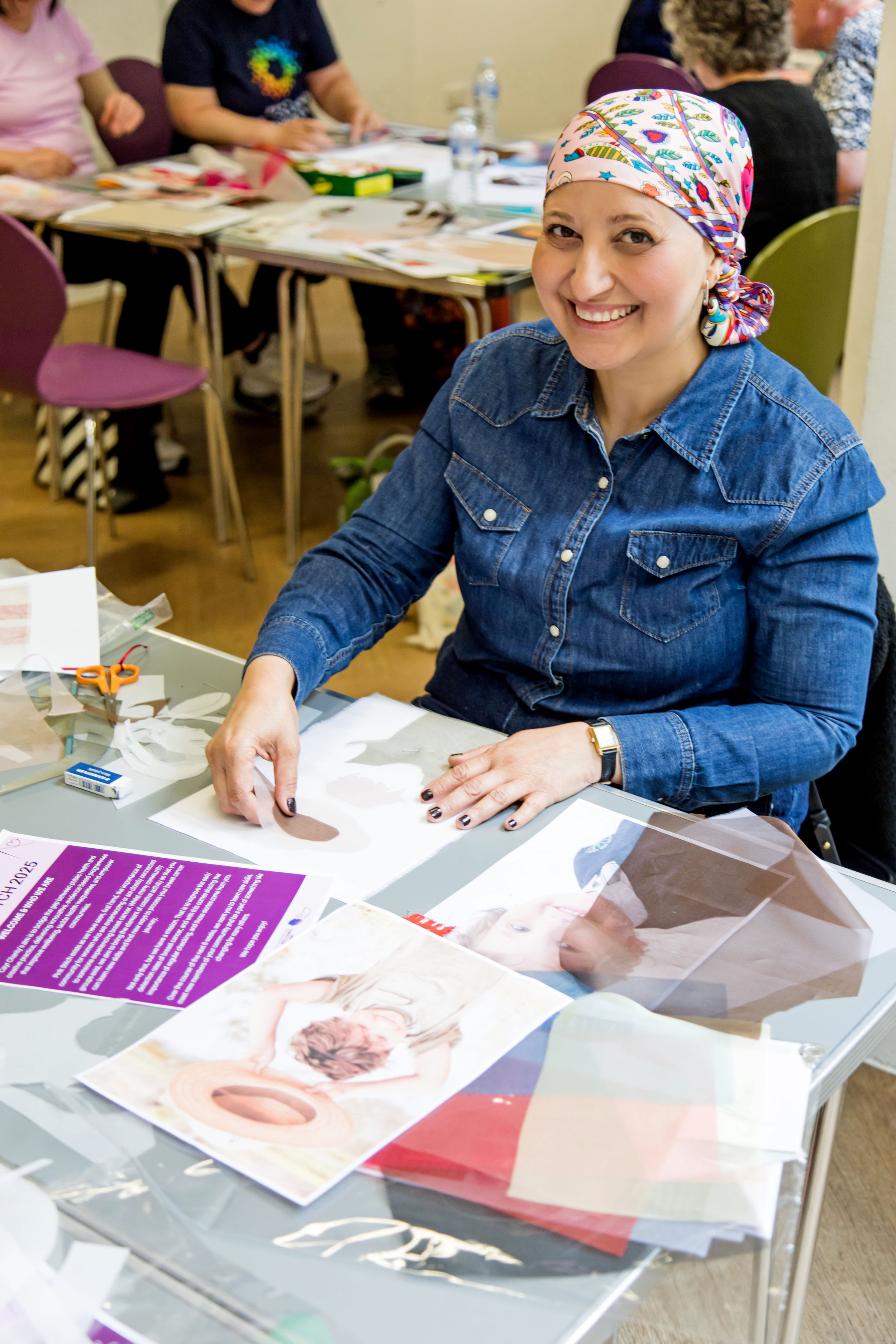 A woman with a colorful bandana and denim shirt smiling at the camera while working on a craft project at a table filled with papers, scissors, and photos, in a group setting.