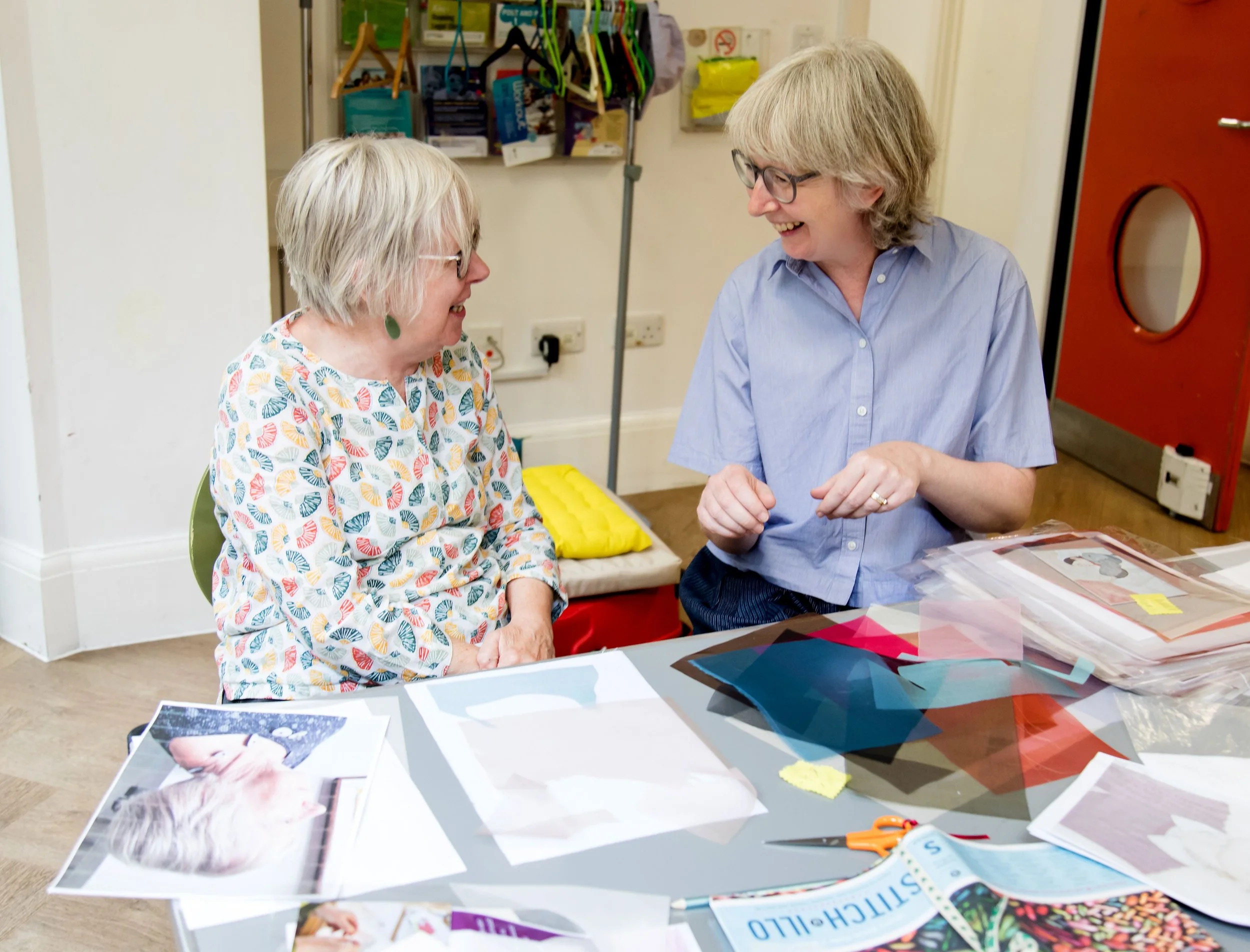 Two women smiling and talking at a table covered with fabric samples, photos, and scissors in a craft or fabric store.