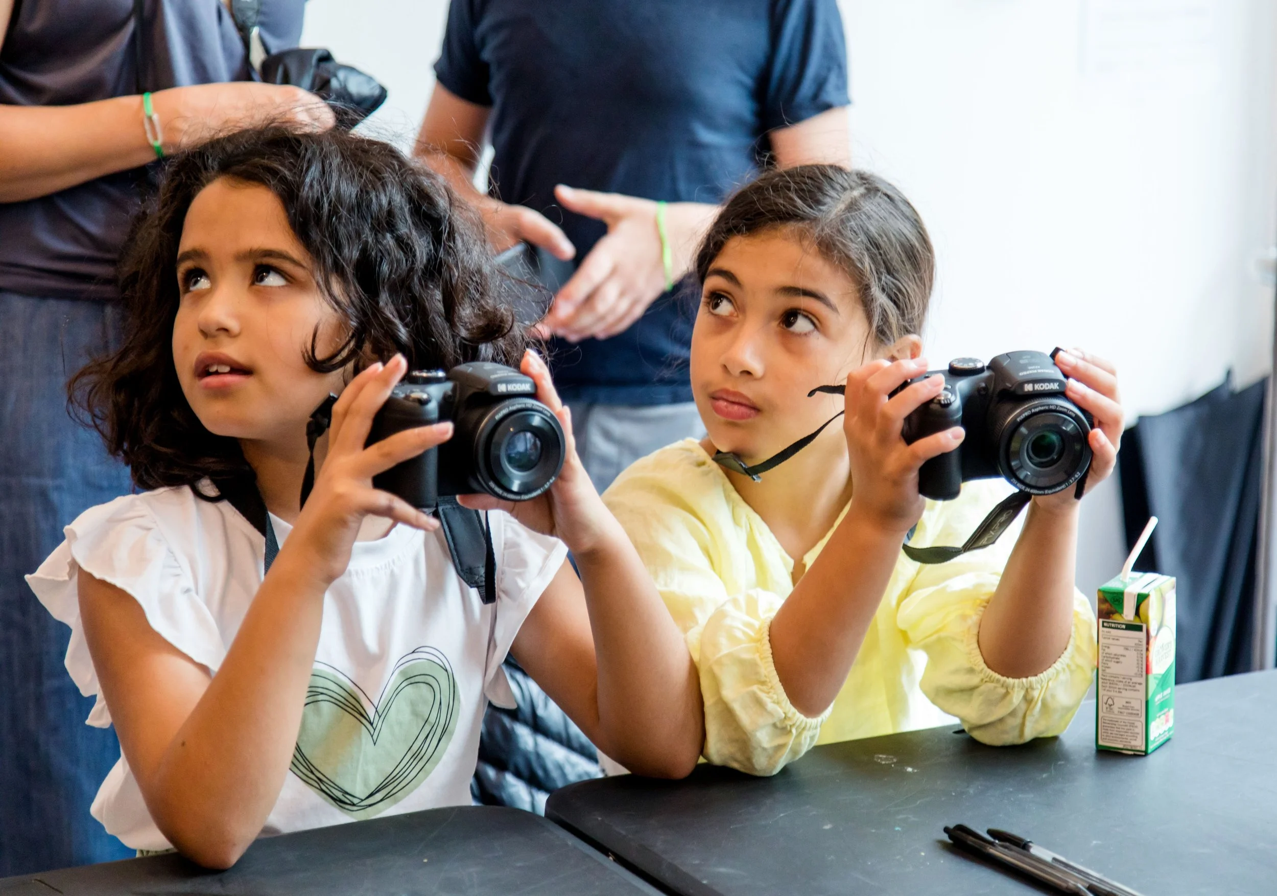 Two young girls sitting at a table holding cameras, looking upward. One girl has dark curly hair and a white shirt with a heart, the other has dark straight hair in a ponytail and a yellow shirt. There are two adults standing behind them, with hands gesturing as if explaining something. A juice box and pens are on the table.