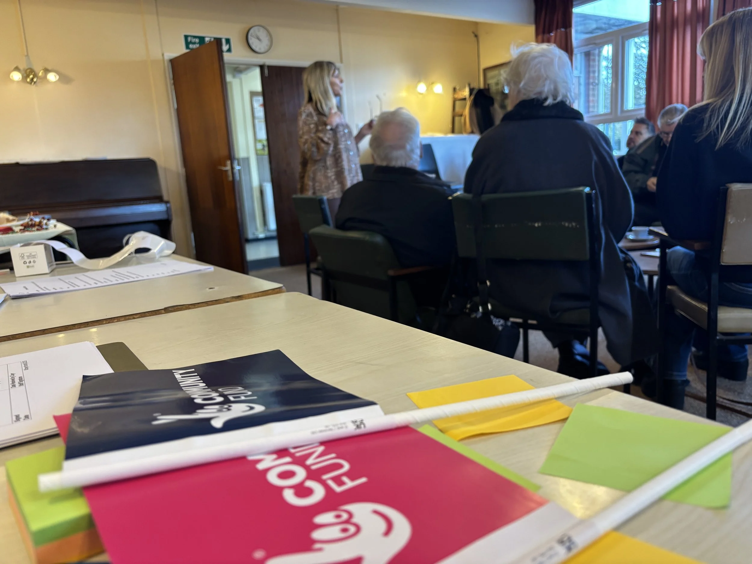 A group of people seated around a table in a room listening to a woman standing and speaking. There are papers, pamphlets, and colorful paper on the table in the foreground. The room has windows with curtains, a piano, and various decorations.