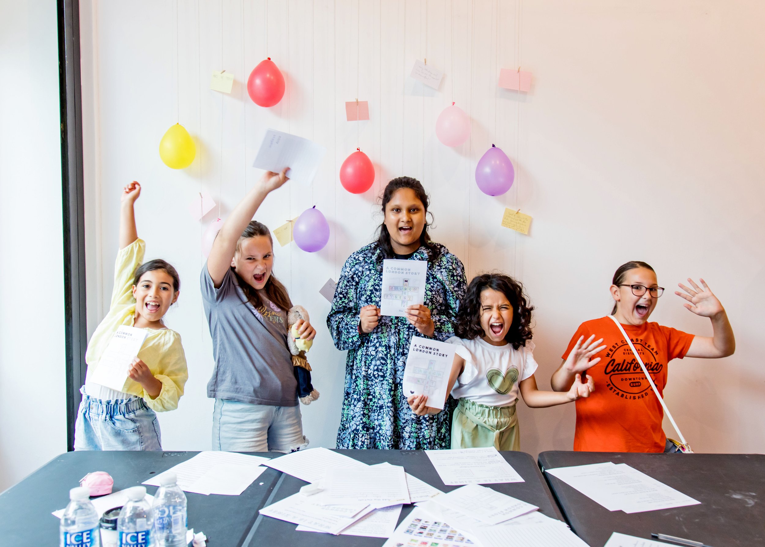 Group of five children celebrating at a table with papers, balloons and notes on the wall behind them