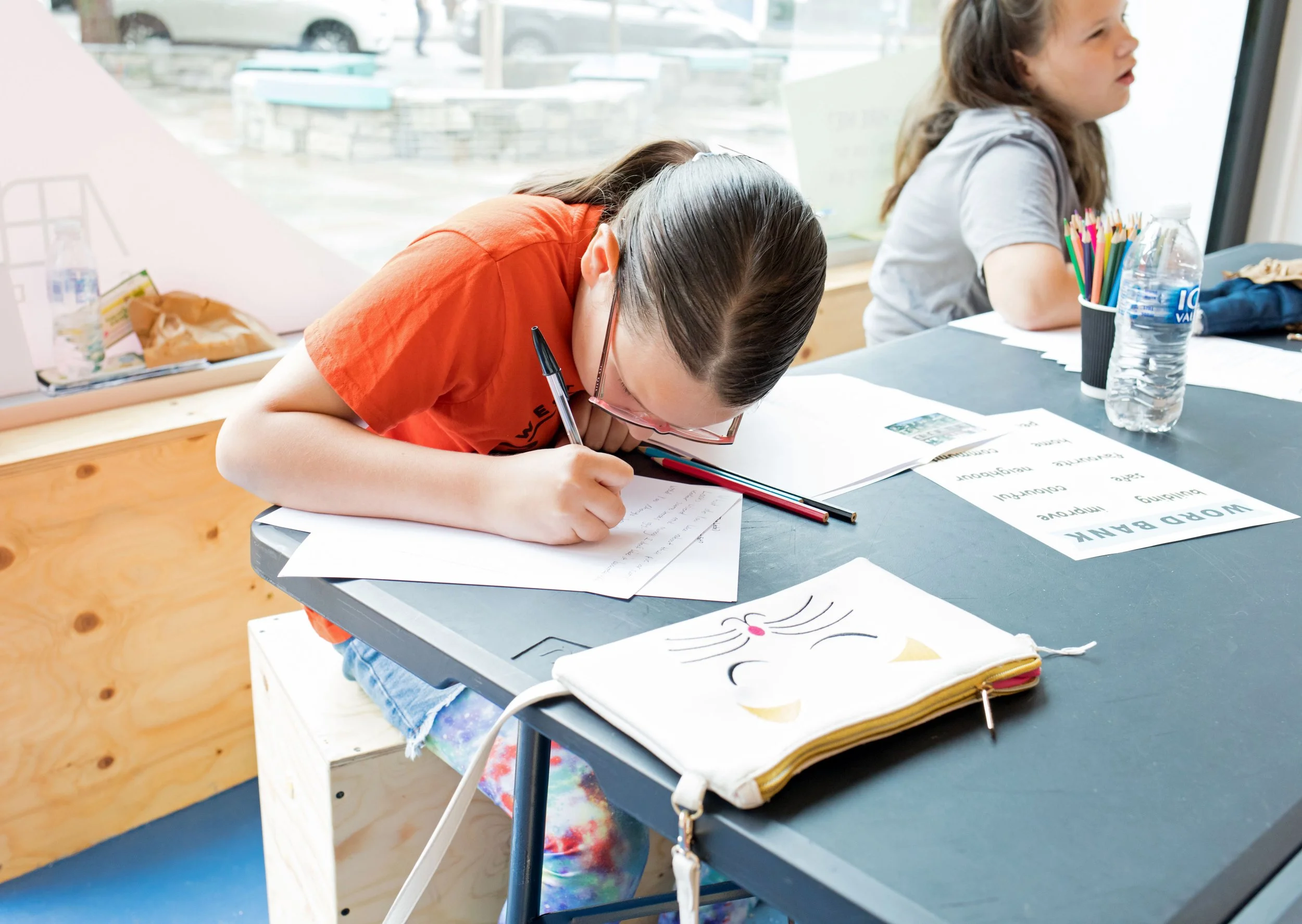A young girl in an orange shirt and glasses is leaning over a table, writing on a piece of paper with a black pen in an indoor classroom setting. In front of her are colorful markers, a cartoon face zippered pouch, and paper with printed words and images. Another girl in gray is sitting at the same table, looking out the window. The table has water bottles and various papers.
