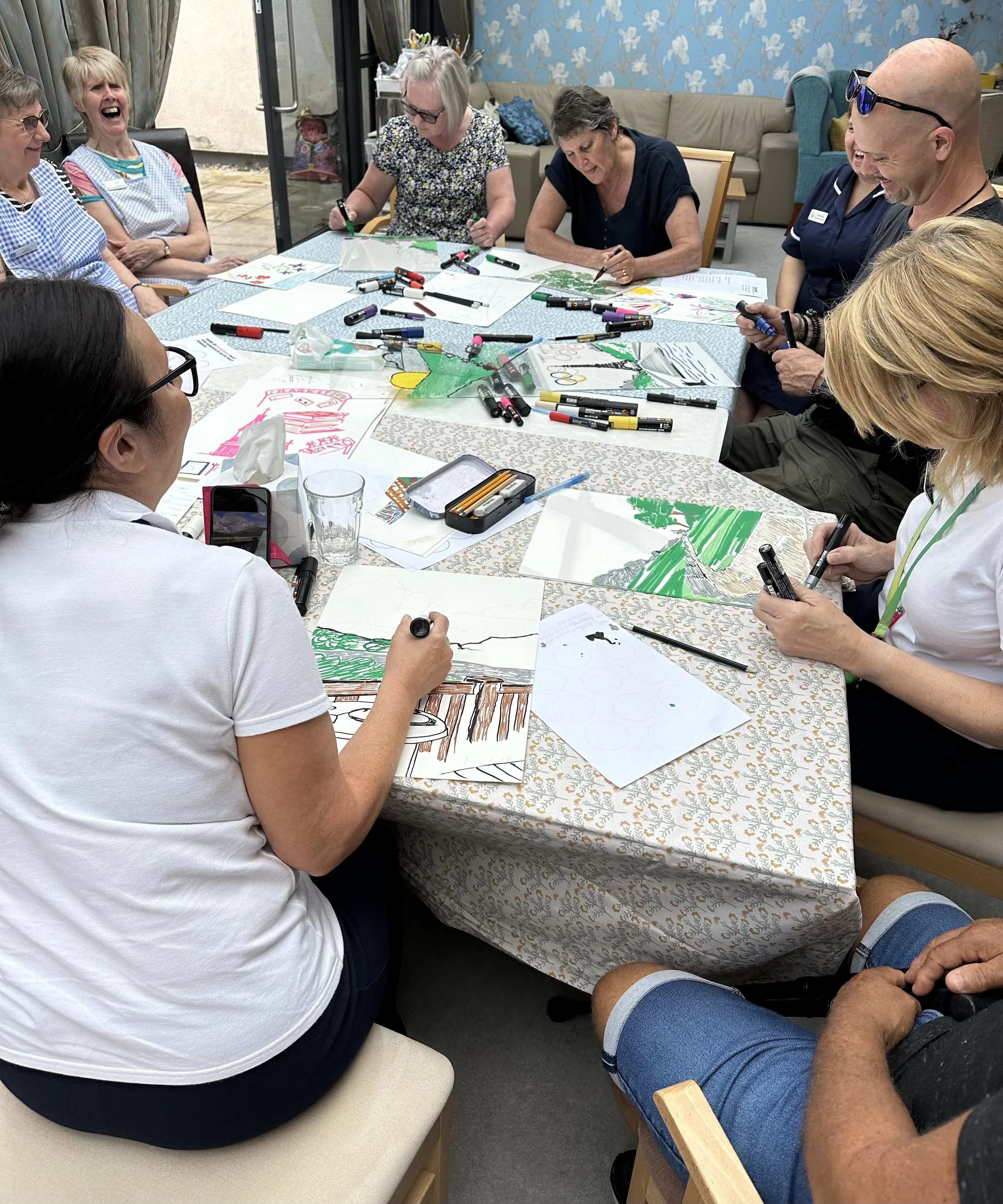 Group of people sitting around a table, coloring and drawing on paper, with art supplies scattered across the table, and some people laughing and smiling.