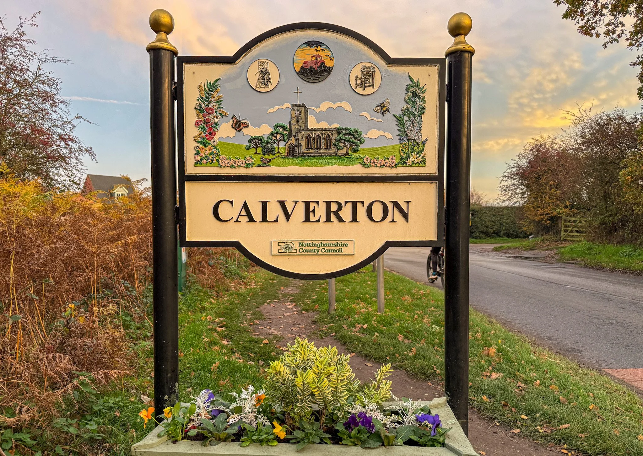 A town entrance sign for Calverton, featuring a church, colorful flowers, and images of a tower, a bridge, and machinery, with a sunset sky in the background.