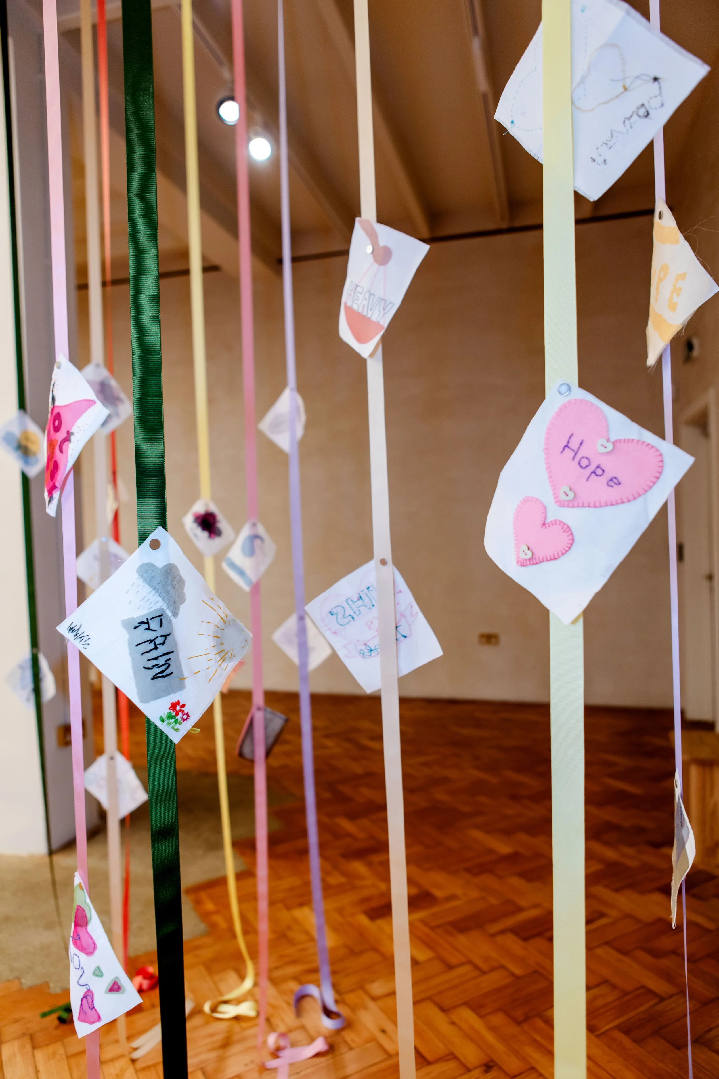 Colorful ribbons hanging vertically with heart-shaped and rectangle-shaped paper notes attached, some with drawings and words like 'Hope' and 'Love'.