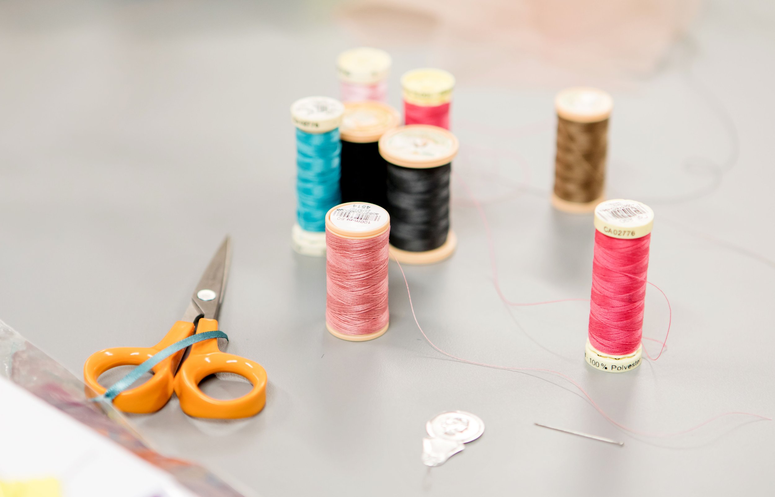 Various spools of sewing thread in different colors, a pair of orange scissors, and a sewing needle on a gray surface.