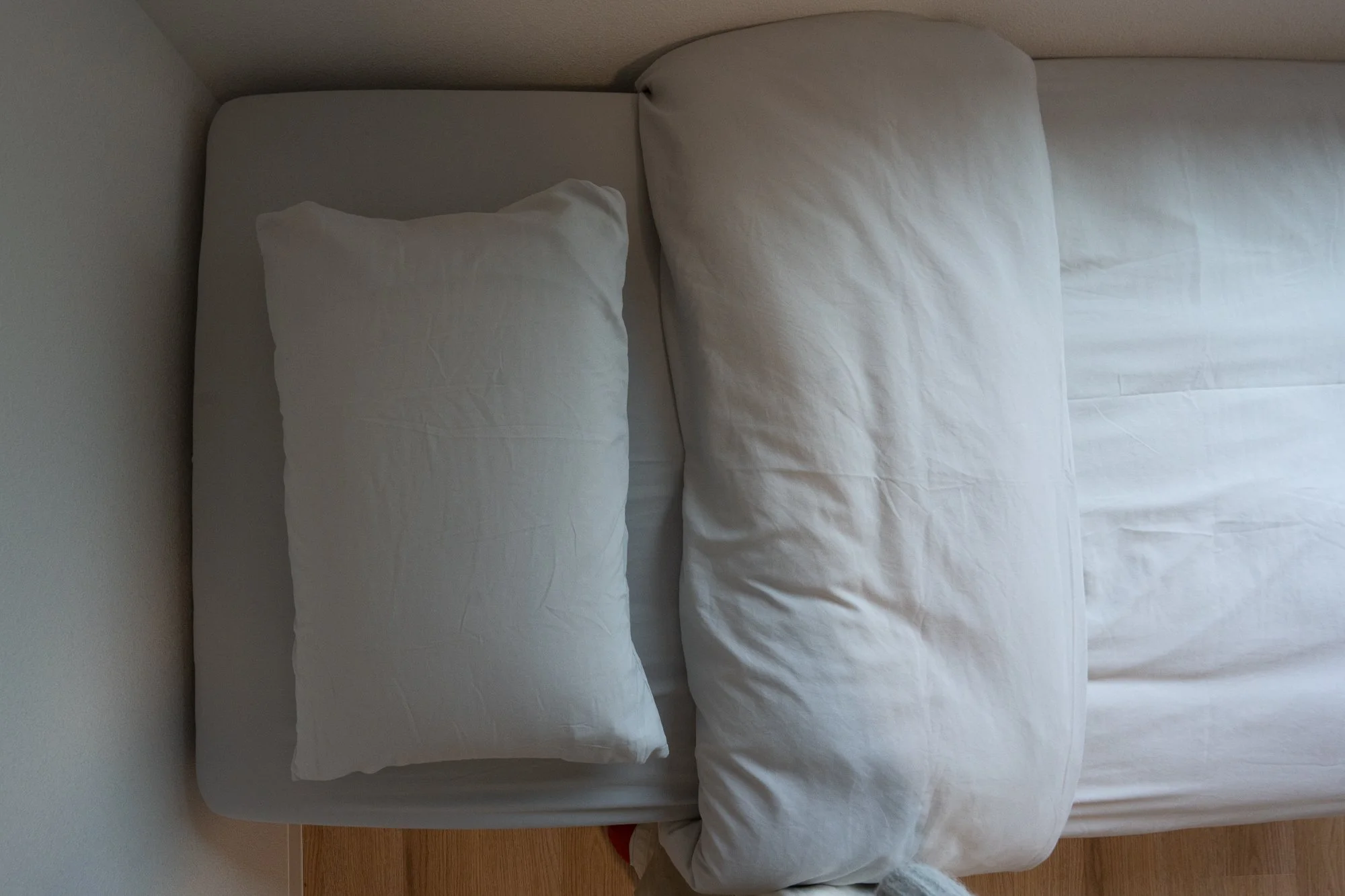 Top view of a bed with white sheets, two pillows, and a folded blanket on a wooden floor.