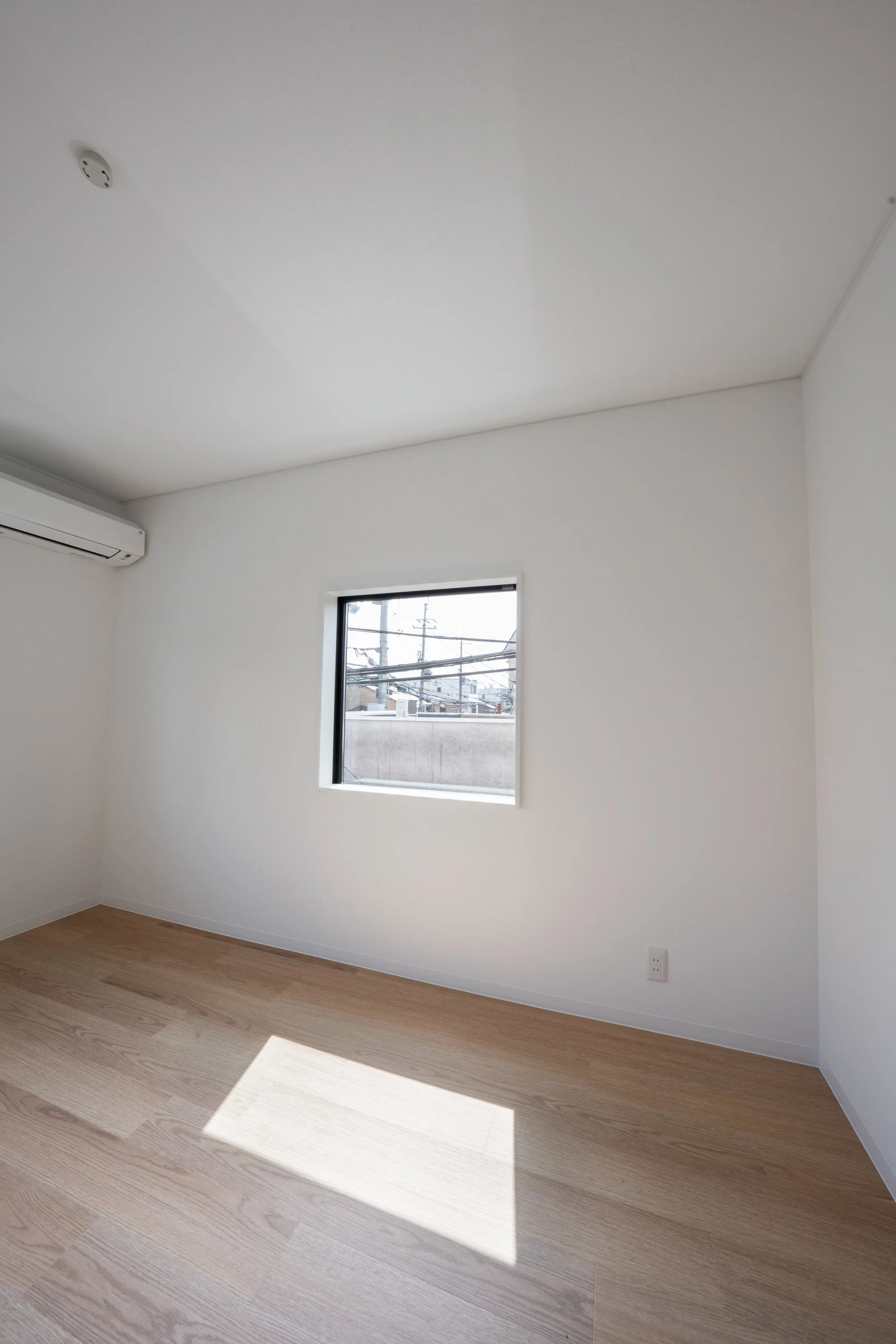 Empty room with white walls, wooden flooring, a small window, and an air conditioning unit on the wall.