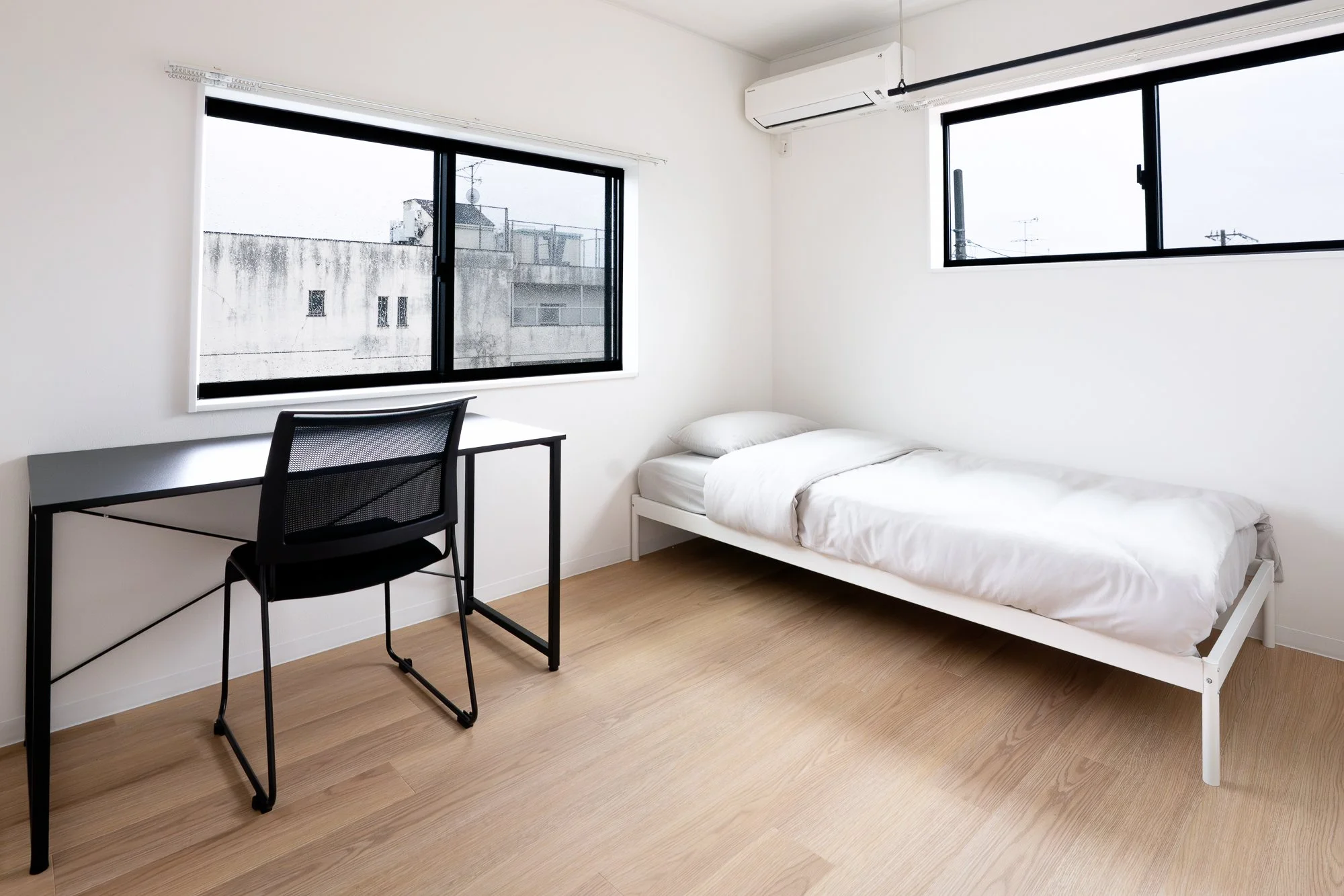 A minimalistic bedroom with a single bed, a desk with a black office chair, white walls, two large windows, and light wood flooring.