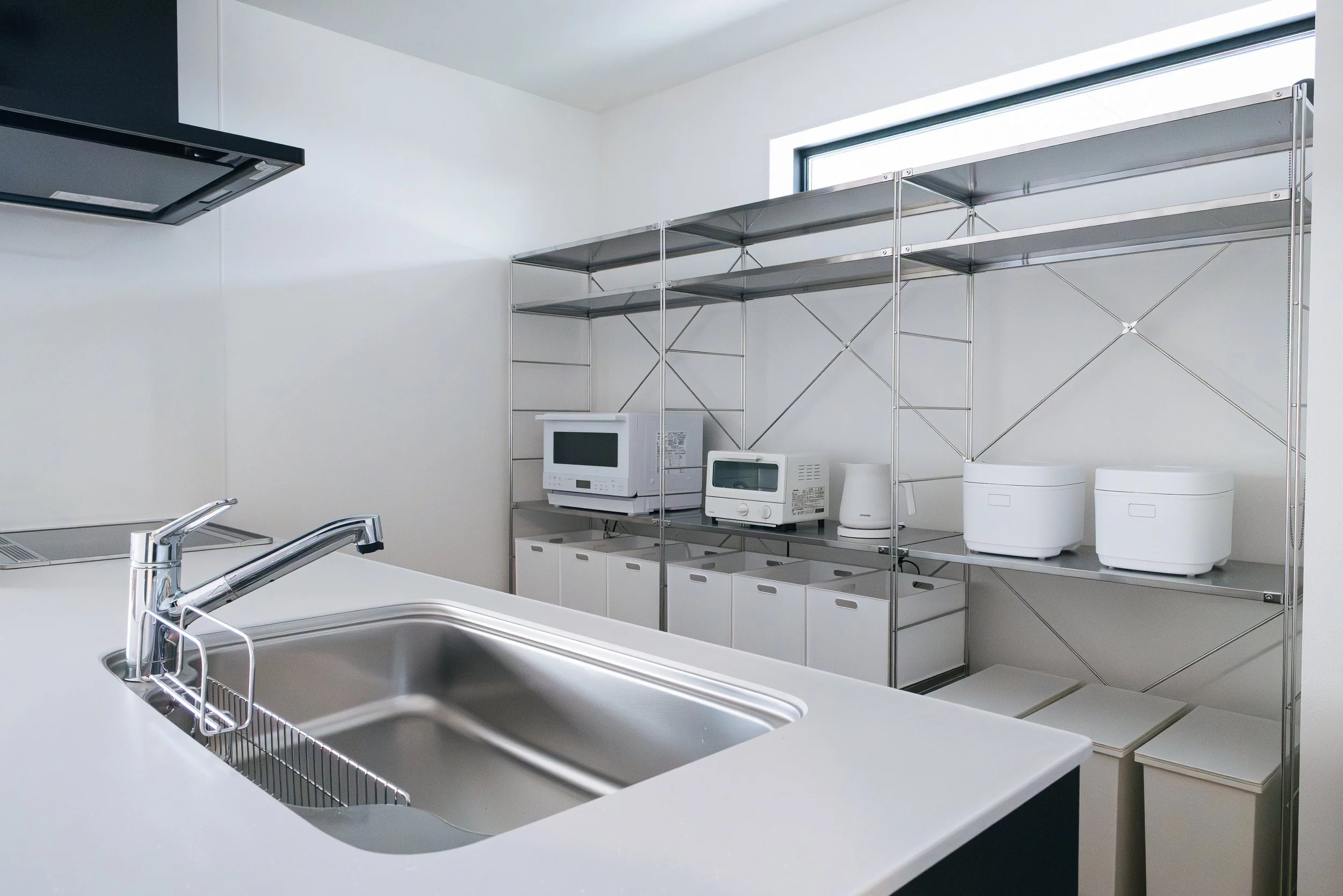 A kitchen with a white countertop, stainless steel sink, and a modern faucet. In the background, metal shelving holds appliances including a microwave, toaster, rice cooker, and food storage containers, with a window providing natural light.