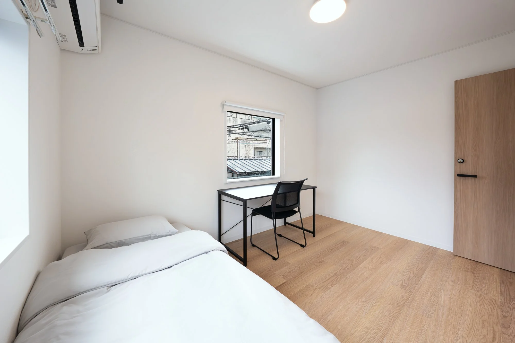 A minimalistic bedroom with a white bed, a black desk under a window, a black chair, wooden flooring, a white ceiling, a circular ceiling light, and an air conditioning unit on the wall.