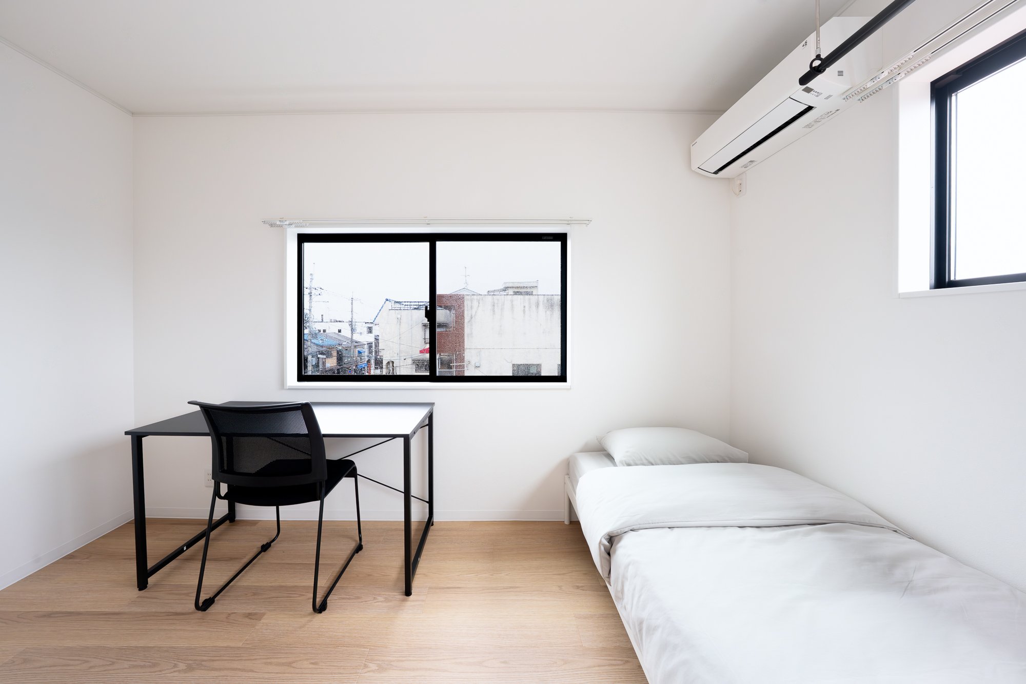 Minimalist room with a white bed, black desk, and black chair, two windows with a city view, and an air conditioning unit.