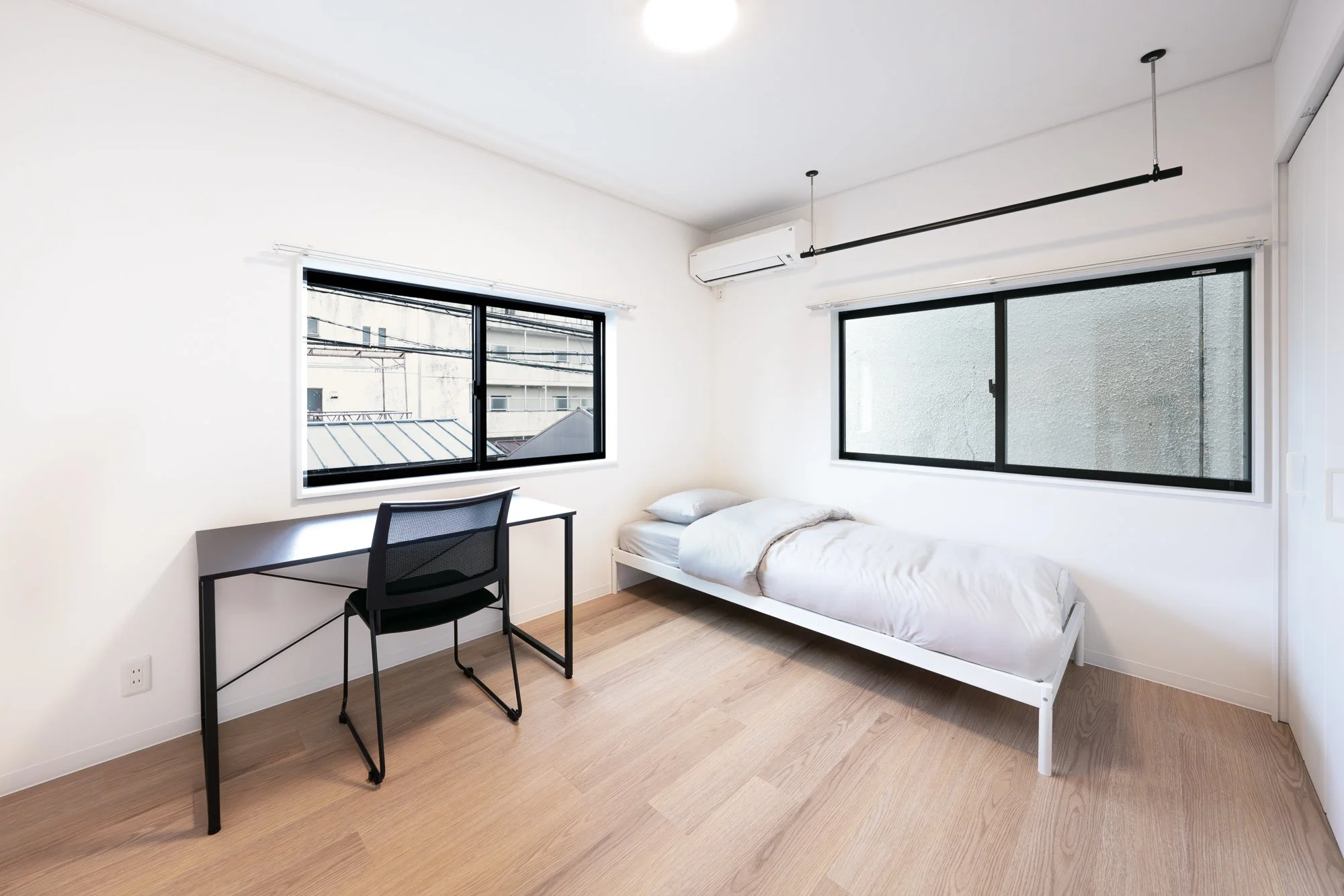 Minimalist bedroom with white walls, a single bed with white bedding, a black desk, a black mesh chair, and two large windows with black frames letting in natural light.