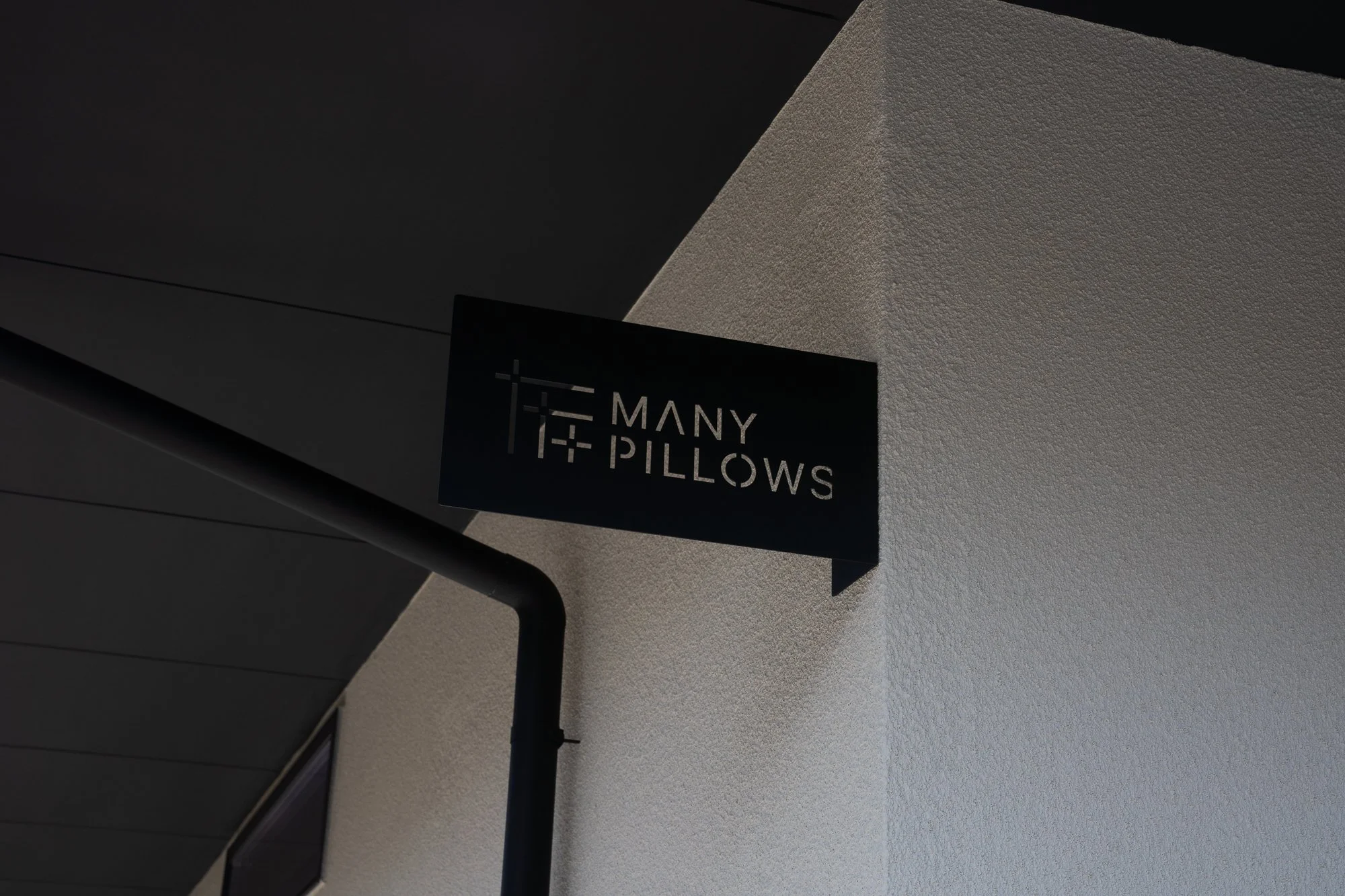A black sign with white text reading 'Many Pillows' is attached to a black metal railing, with part of a textured beige wall visible in the background.