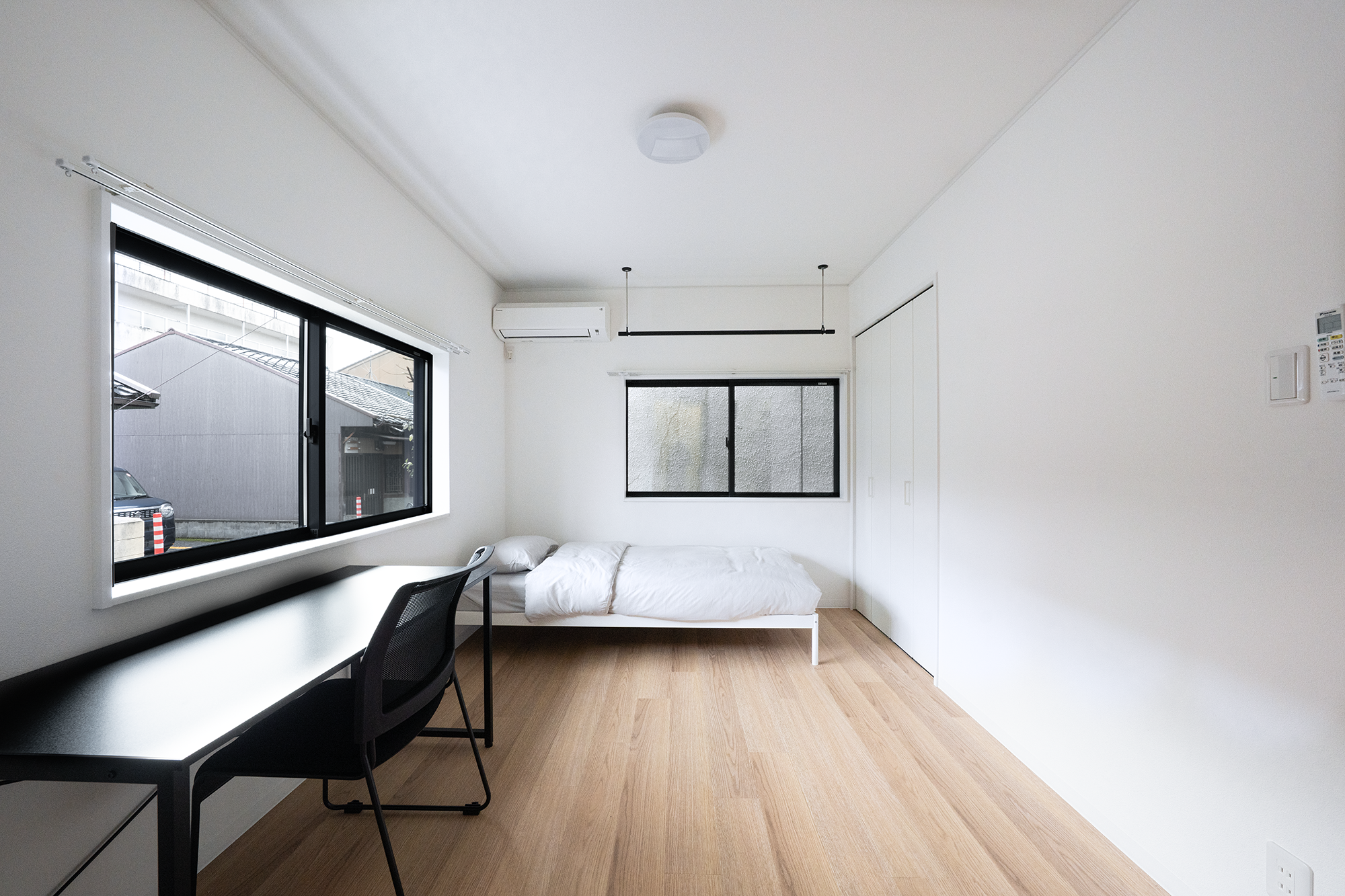 A minimalist, bright bedroom with light wood flooring, a white bed with gray pillows, black-framed windows, a black desk with a chair, and an air conditioning unit on the wall.