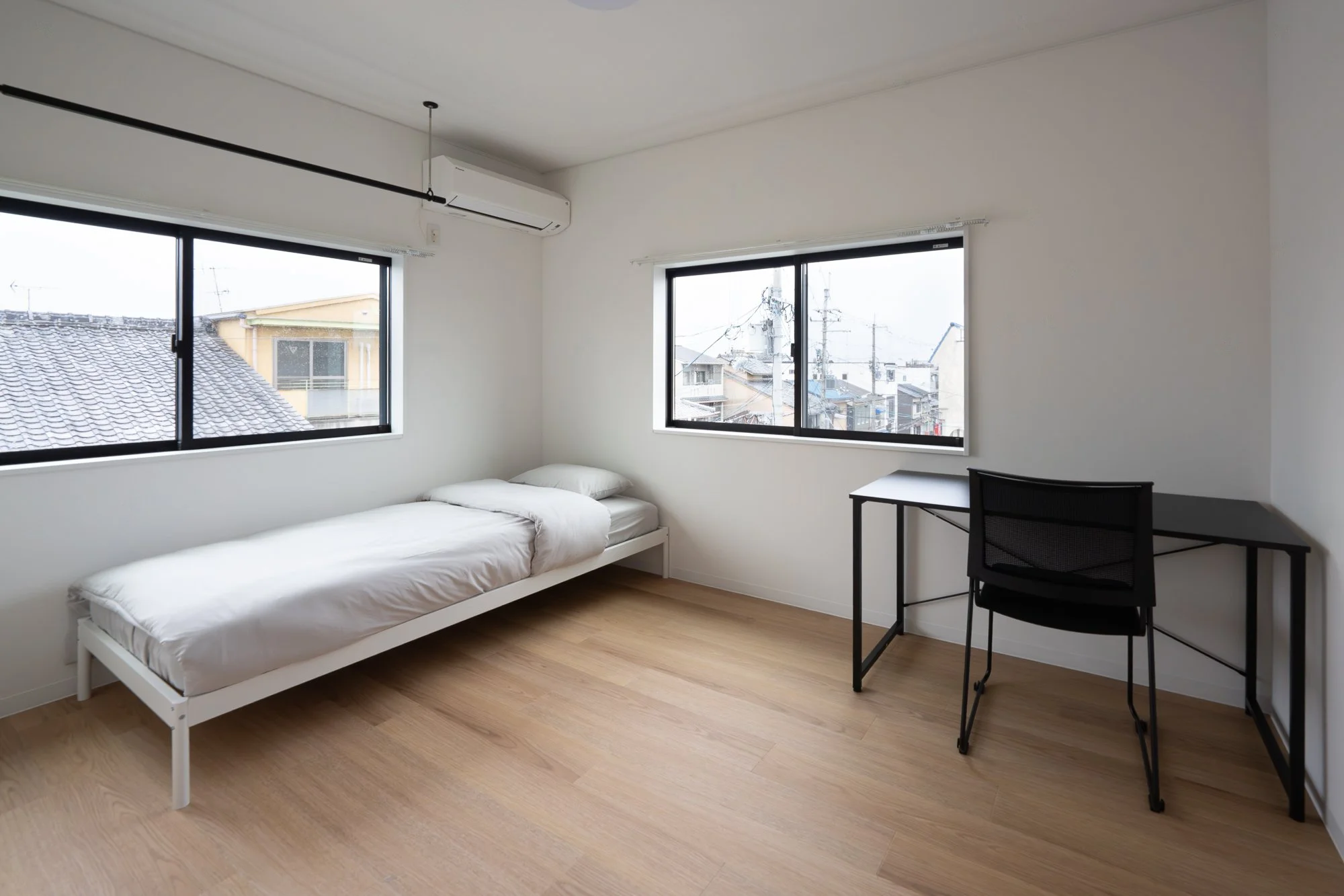A minimalist bedroom with a single bed, white bedding, and a black desk and chair near two windows showing a cityscape with power lines and rooftops.