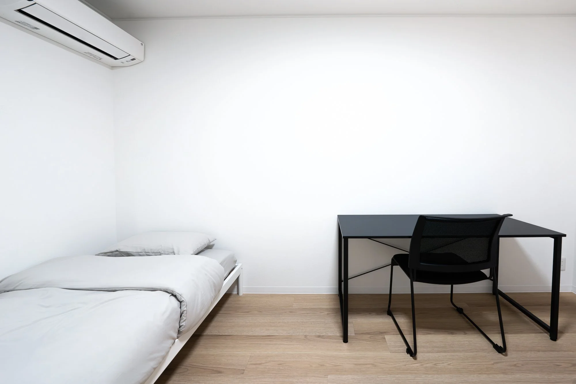 Minimalist room with a white bed, pillow, black desk, and black chair, with white walls, wooden floor, and a wall-mounted air conditioner.