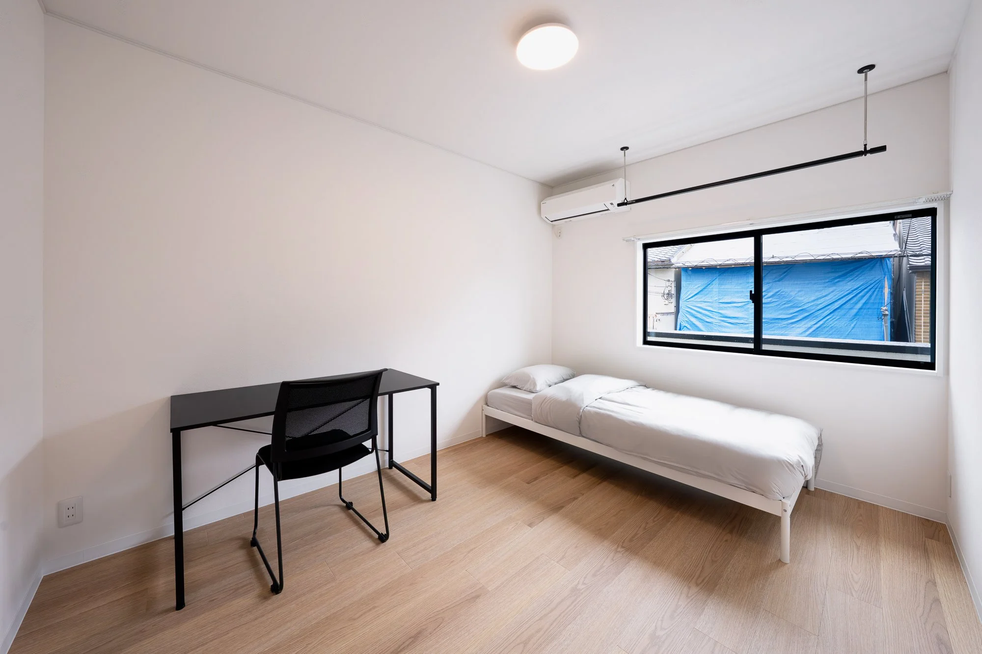 A minimalist bedroom with a single bed, a black desk, and a black chair. There is a large window with a blue outdoor tarp visible outside. The room has white walls and light wood flooring, with a ceiling light and an air conditioning unit.