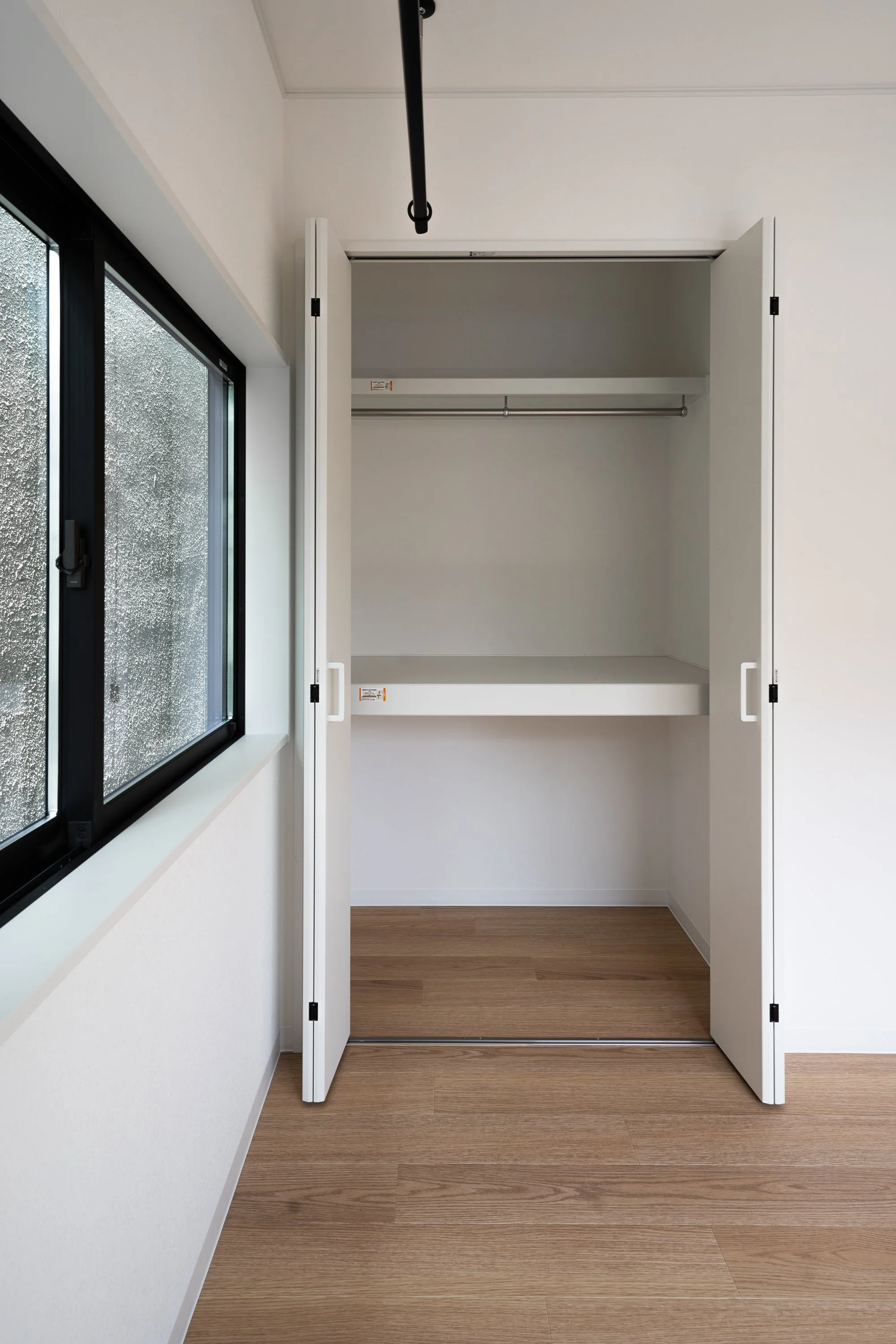 Empty white closet with two doors, a shelf, and a hanging rod, next to a window with raindrops on the glass.