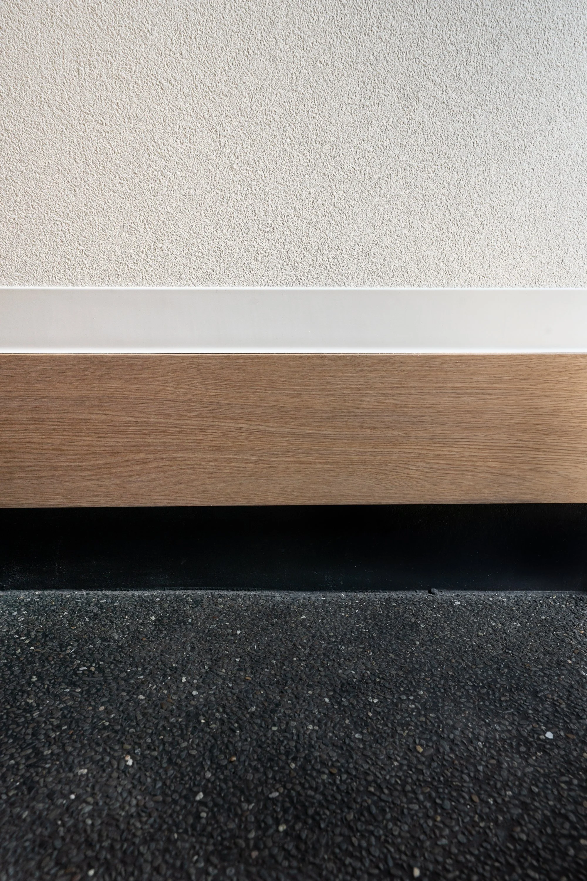 Close-up of a wall baseboard, with a textured white wall above, a wooden strip in the middle, and a black textured flooring below.