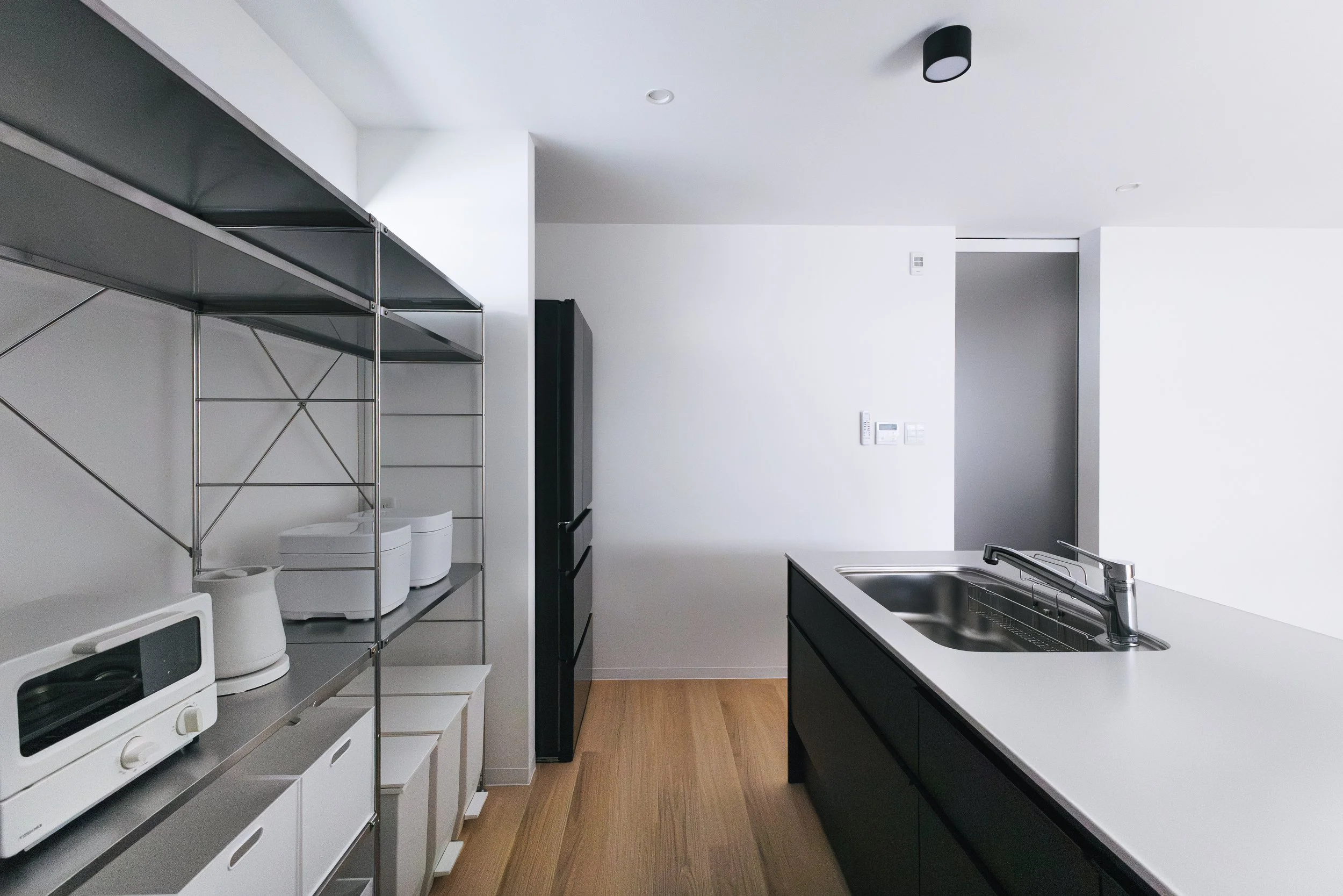 Modern kitchen with white walls, black cabinetry, a stainless steel sink, and minimal decor. Metal shelving unit with small appliances and storage drawers on the left, and a black refrigerator in the background. Light wood flooring.