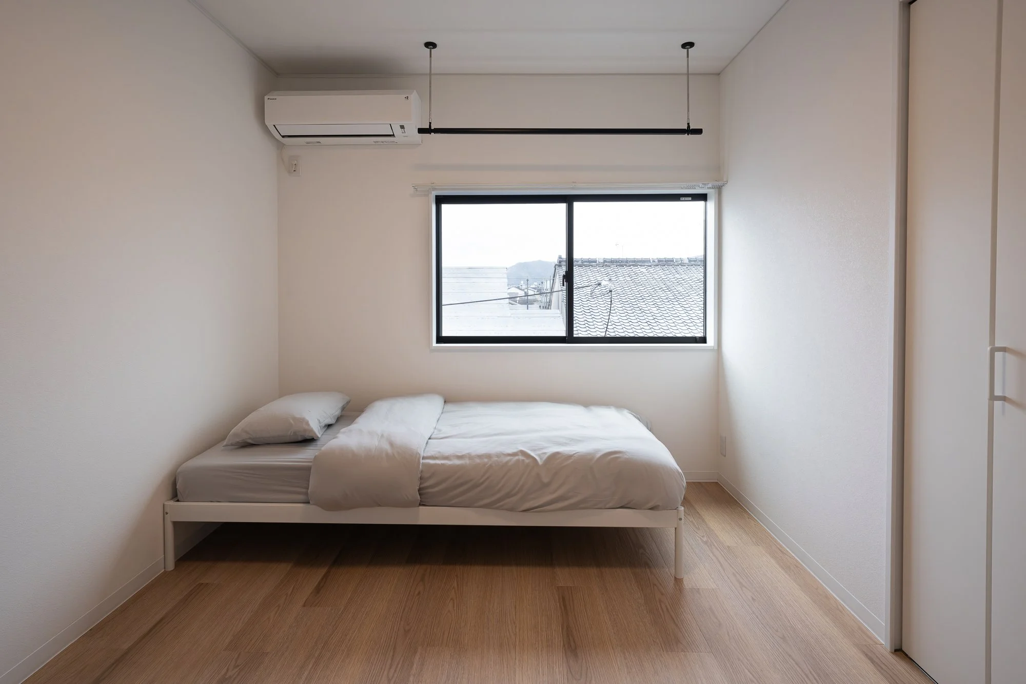 Minimalist bedroom with white bed, white pillows, beige duvet, wooden flooring, window showing rooftops and a distant mountain, air conditioning unit, and a ceiling-mounted hanging rod.