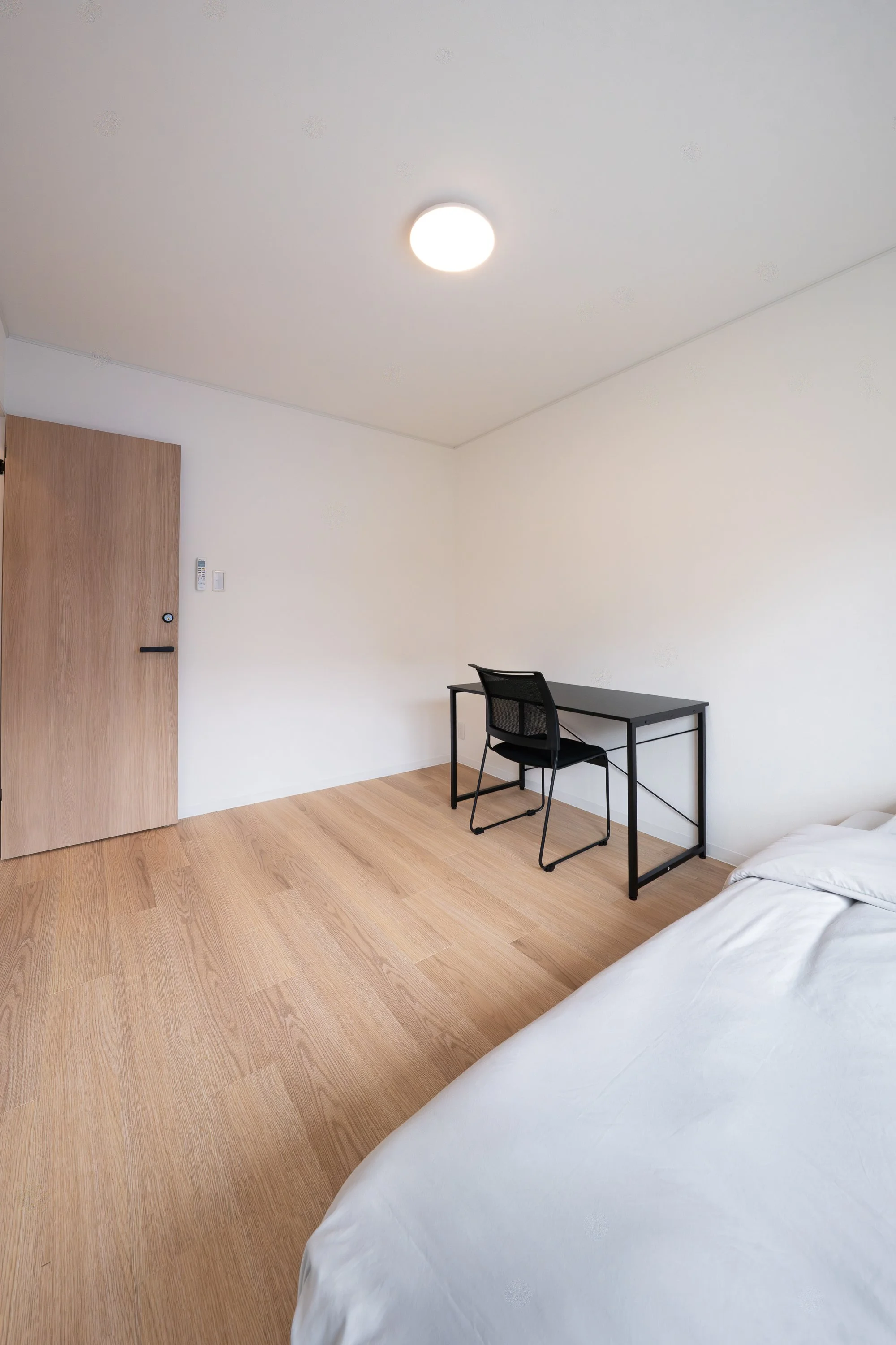Minimalist hotel room with a bed, a wooden door, a small desk with a black chair, and a ceiling light