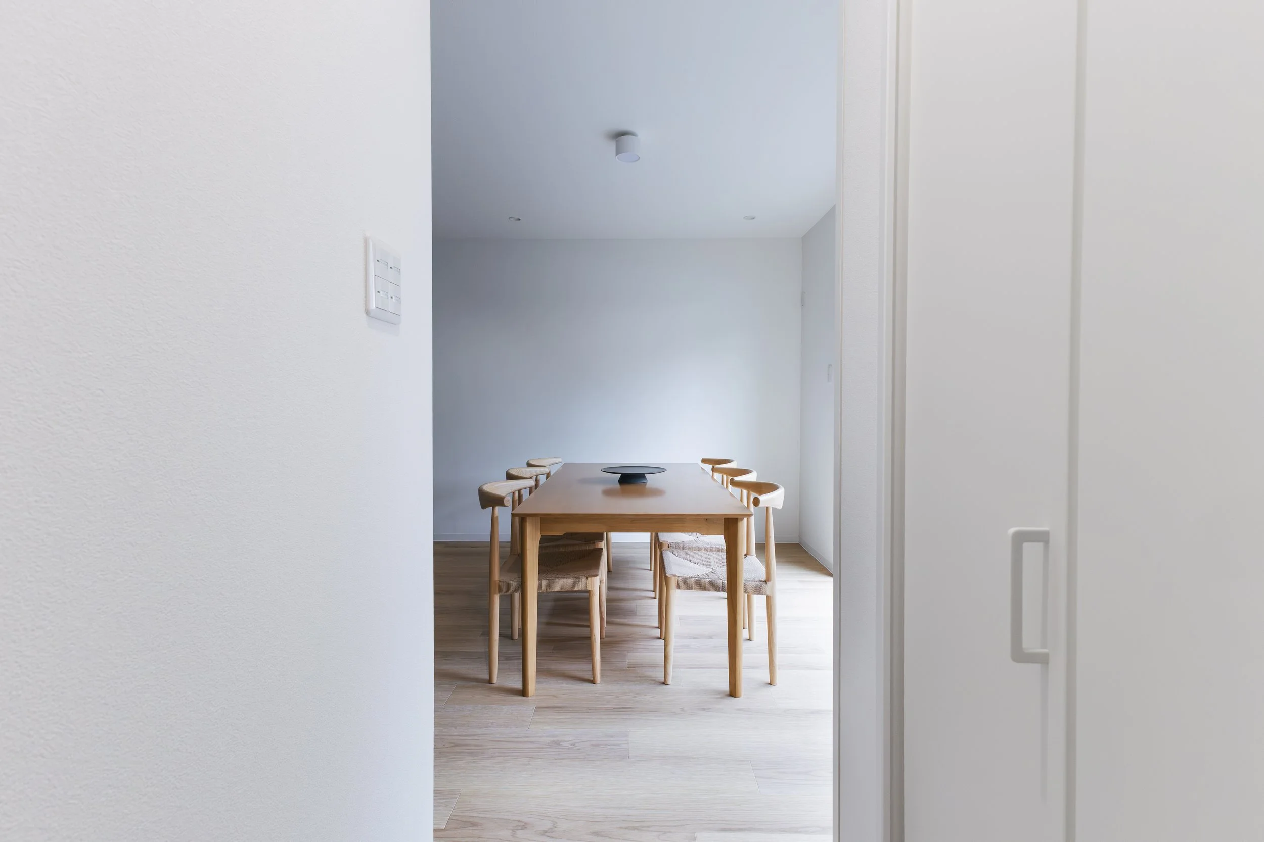 A dining area visible through an open door with white walls, a wooden dining table, and six matching wooden chairs with woven seats. The room has light-colored wooden flooring, and the ceiling has a modern, small cylindrical light fixture and recesse