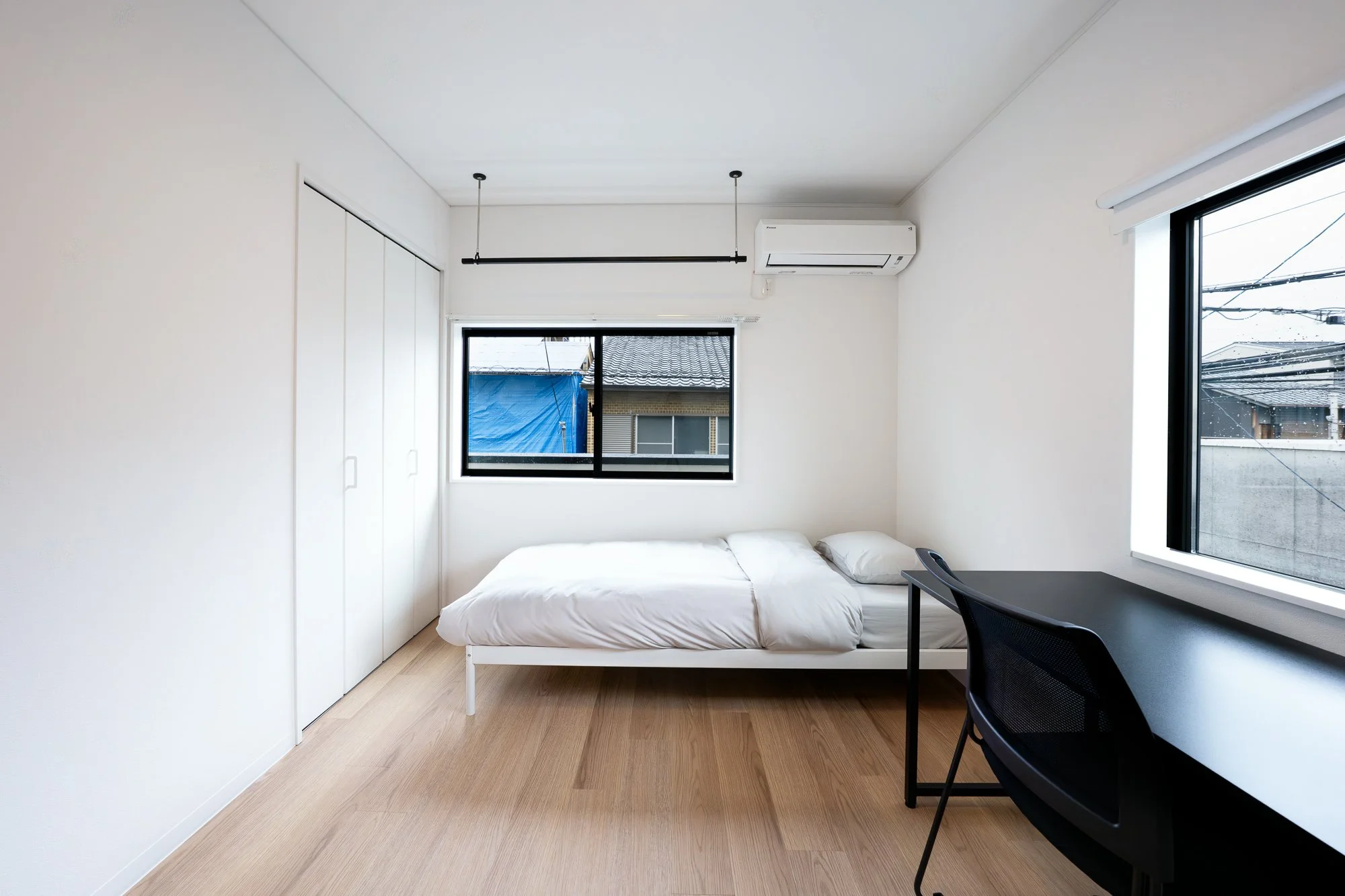 A minimalist bedroom with white walls and light wooden flooring. It features a single bed with white bedding, a black desk with a black chair near a window, a closet with white sliding doors, and windows showing neighboring buildings and power lines.