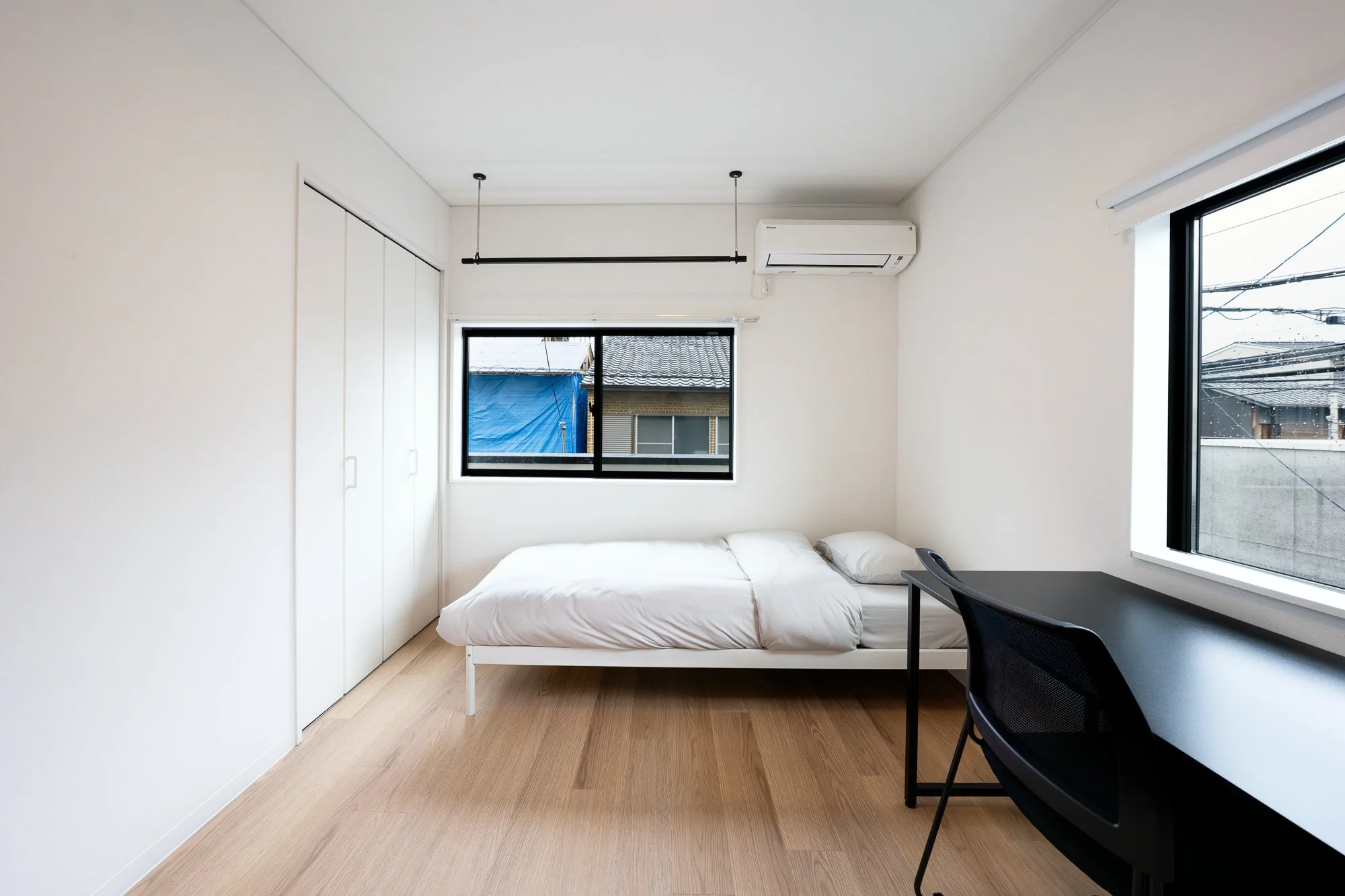Minimalist bedroom with white walls, a bed with white bedding, a black desk with a black chair, and two windows showing neighboring houses and power lines.