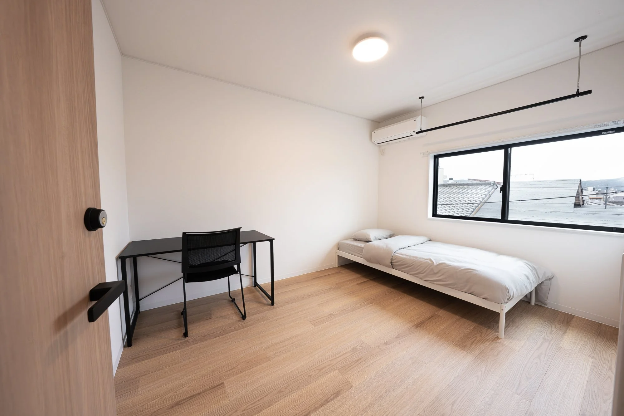 A minimalistic bedroom with a bed near a large window, a black desk with a matching chair, and wood flooring.