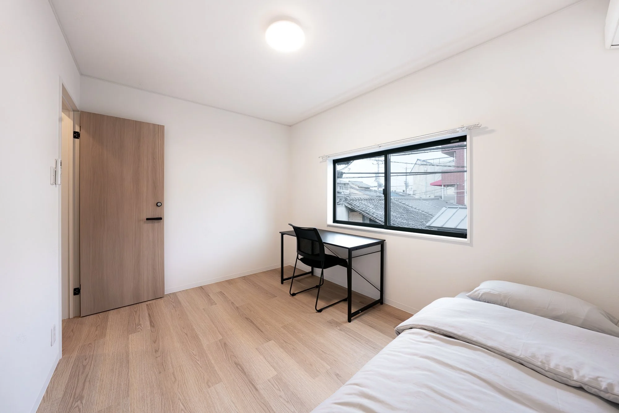 A minimalist bedroom with white walls, light wood flooring, a bed with white sheets, a black desk and chair by a large window, and a closed wooden door.