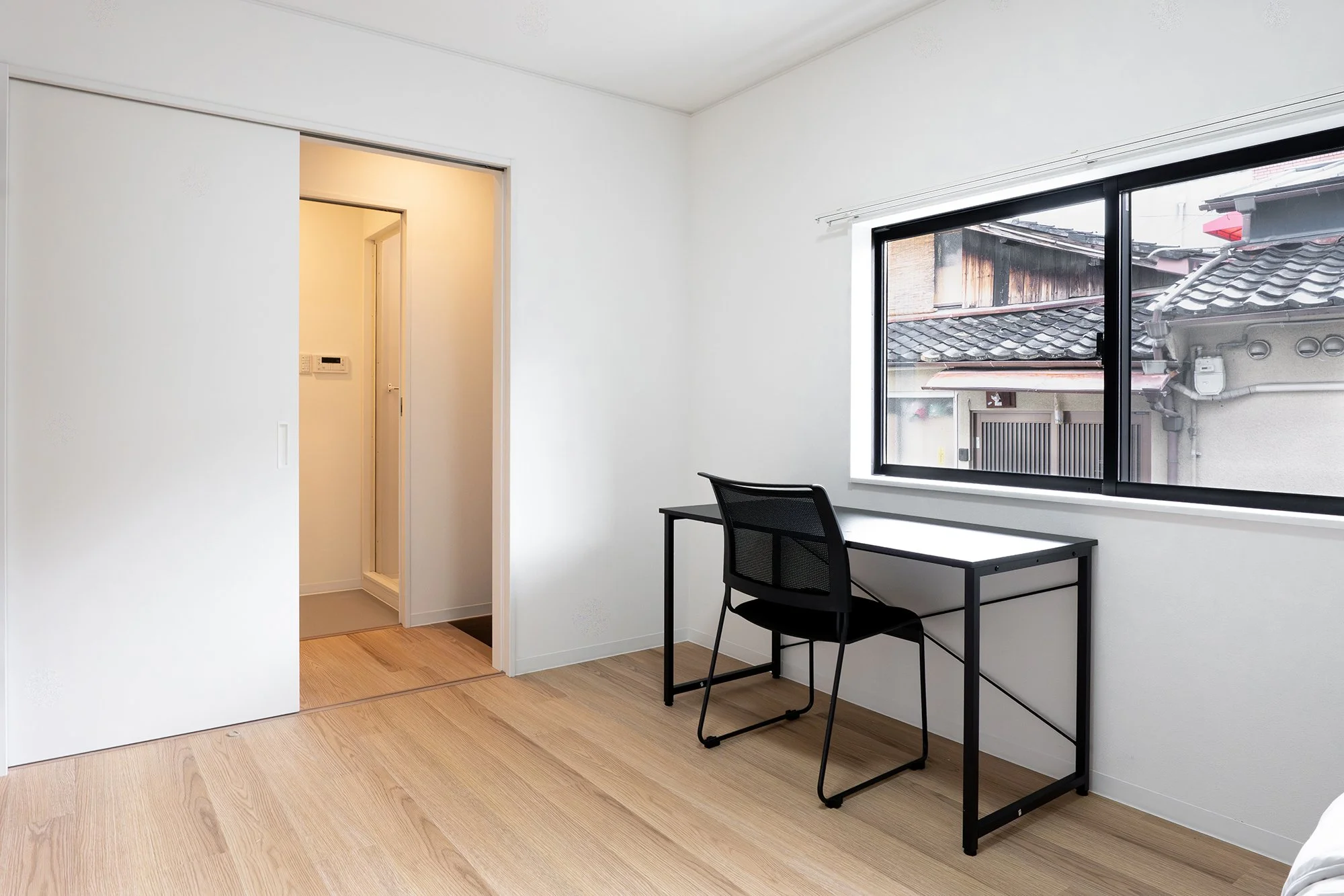 A minimalist room with white walls, wooden floor, black desk, and black chair. Large window showing neighboring buildings with traditional tiled roofs.