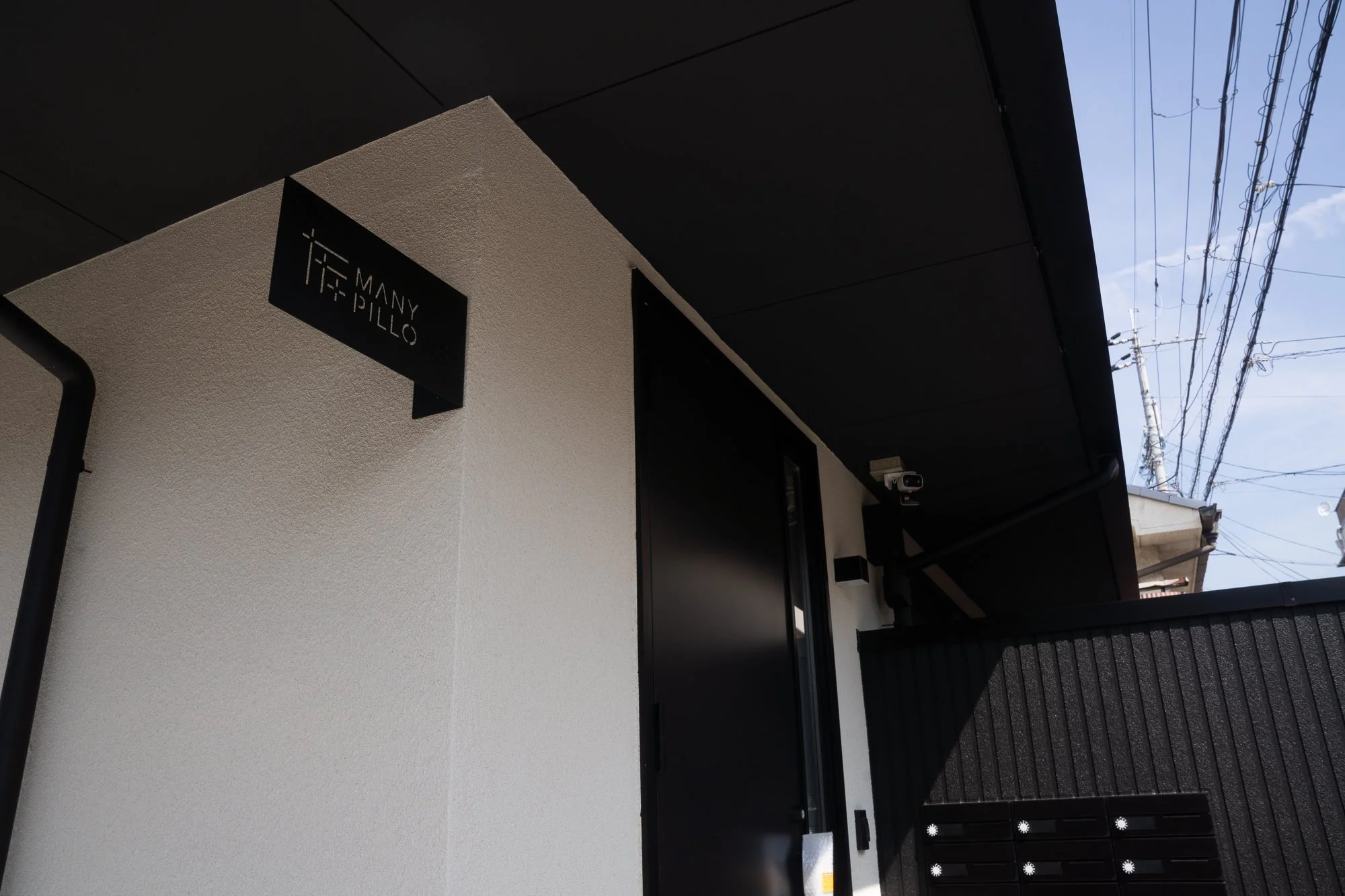 Close-up of the exterior of a modern building with a black sign reading 'Many Pillo', a black door, and a black mailbox, with power lines in the background.
