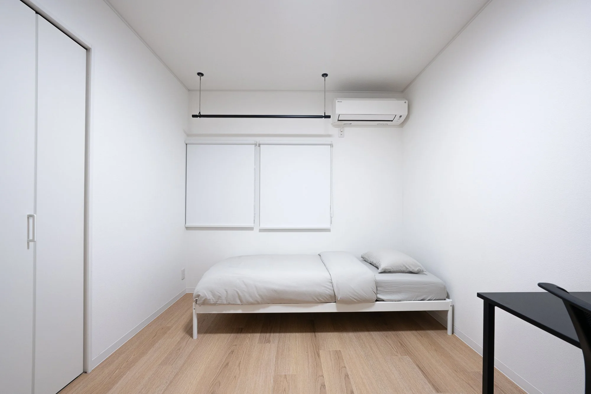 A minimalistic bedroom with a white bed frame, white bedding, and pillows, two windows with blinds, an air conditioning unit, and part of a black desk with a chair.